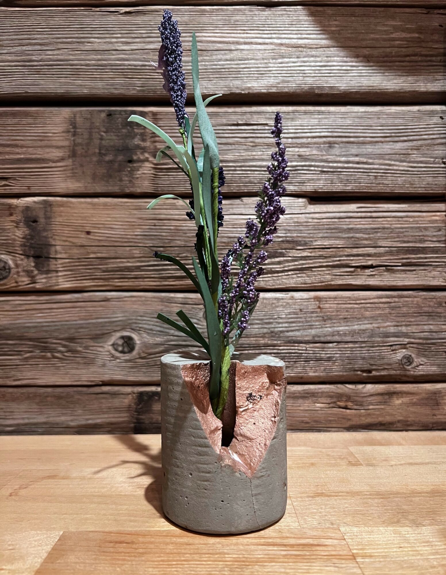 Lavender flowers in a cracked gray ceramic vase on a wooden surface with a wooden wall background.