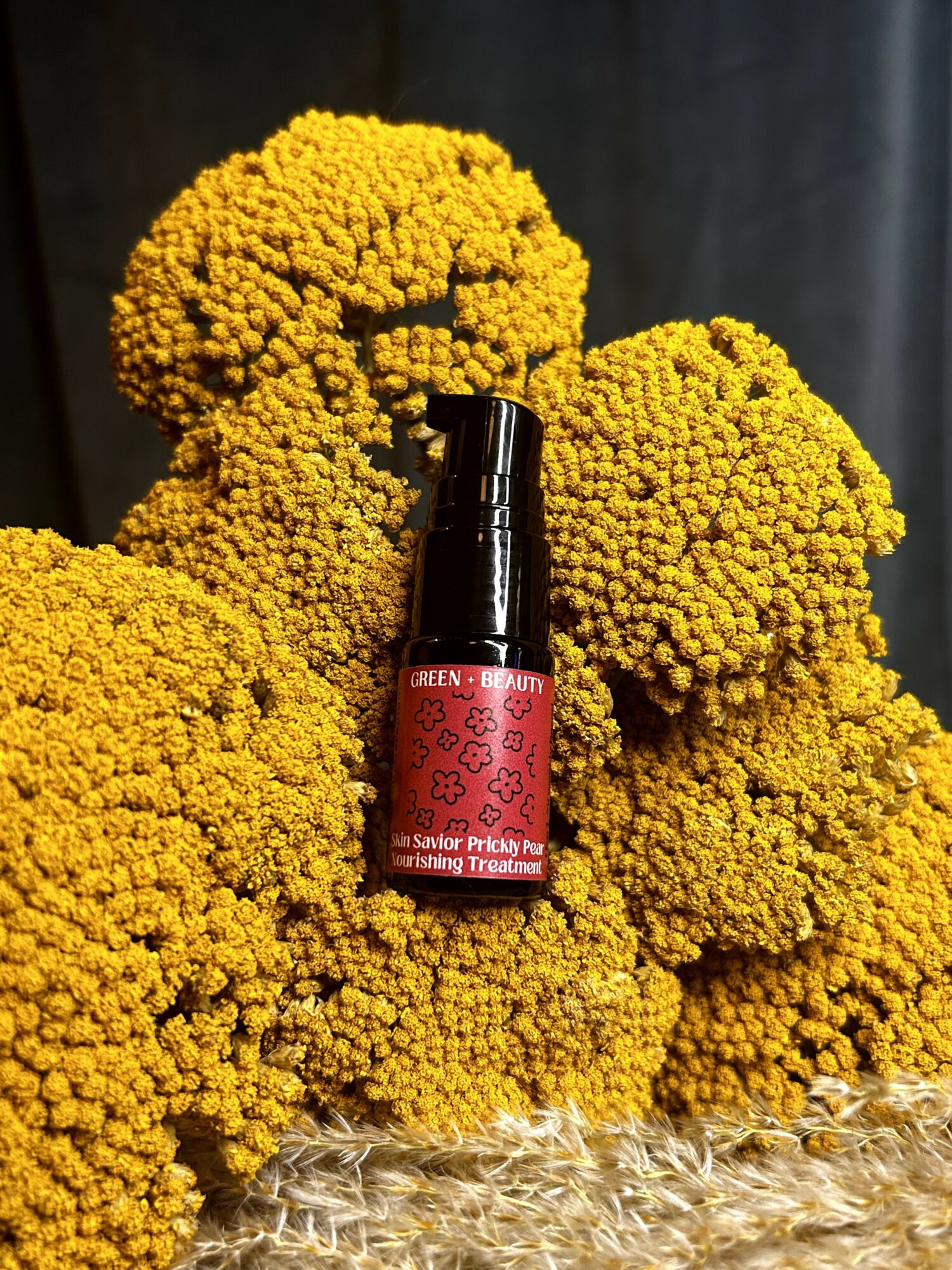 Small red bottle with black cap placed among yellow flowers, dark background, and a textured surface.