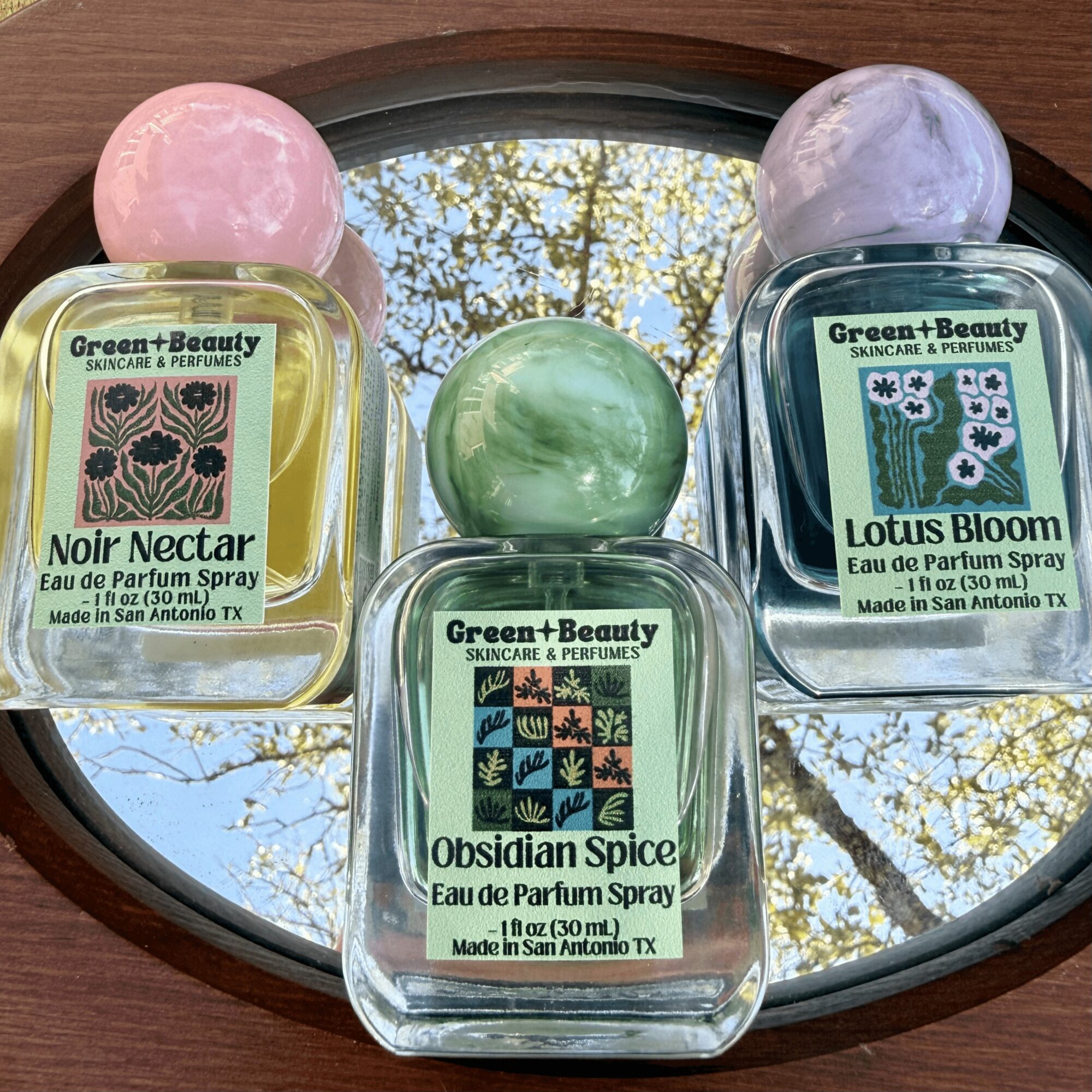 Three perfume bottles with colorful bath bombs on a mirror, reflecting trees and sky. Labels read 'Noir Nectar,' 'Obsidian Spice,' and 'Lotus Bloom.'