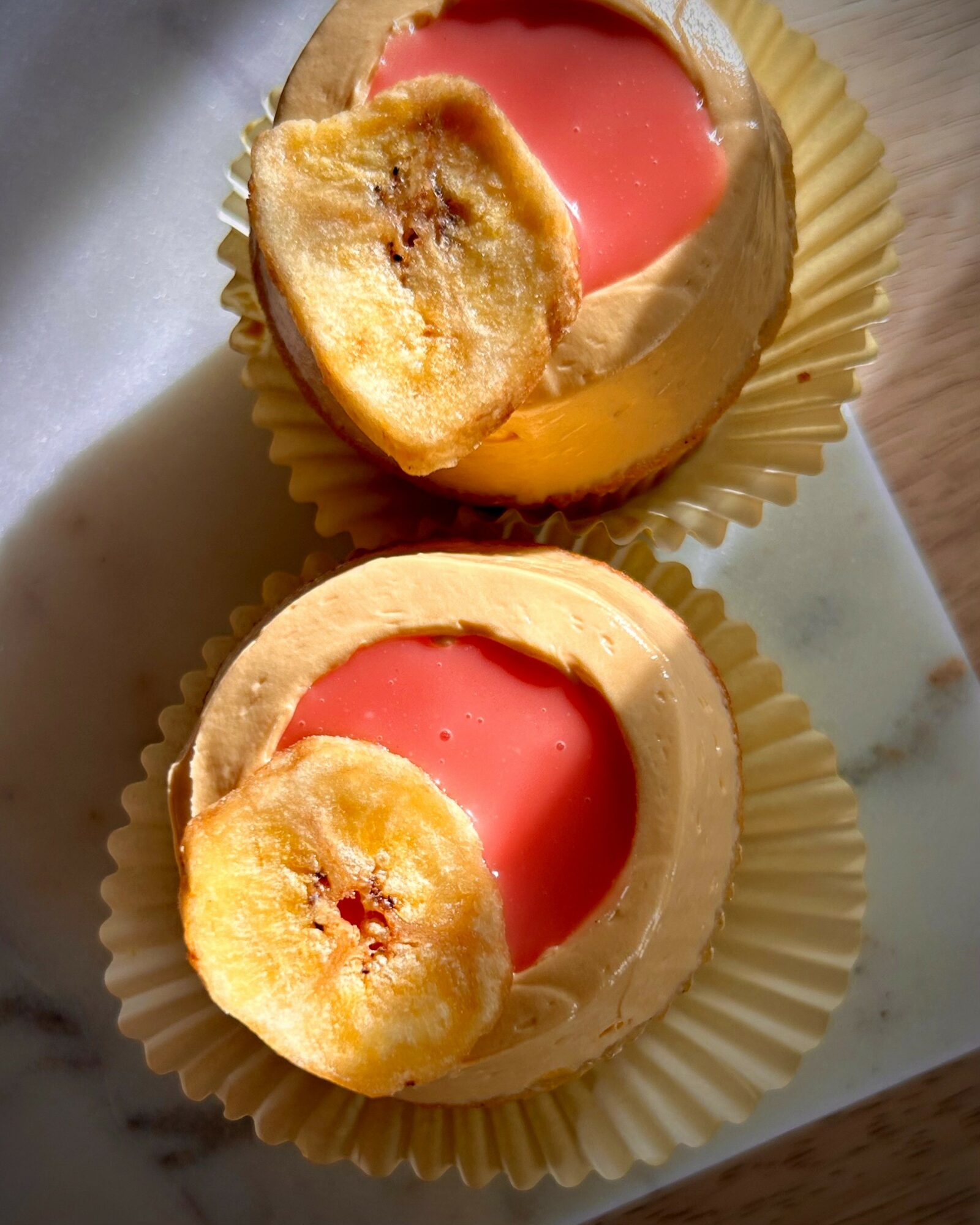 Two cupcakes with pink frosting, topped with banana slices, in yellow paper cups on a white surface.
