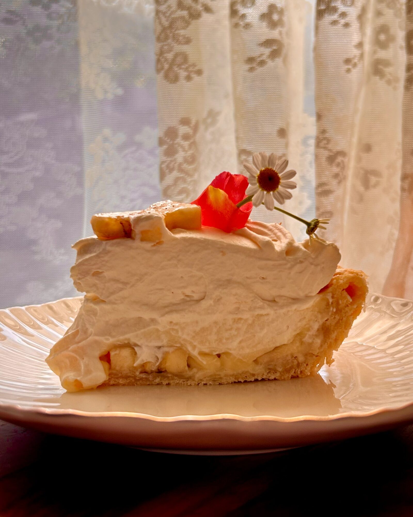 Slice of cake with white frosting, decorated with a small flower and fruit, on a plate in front of a lace curtain.