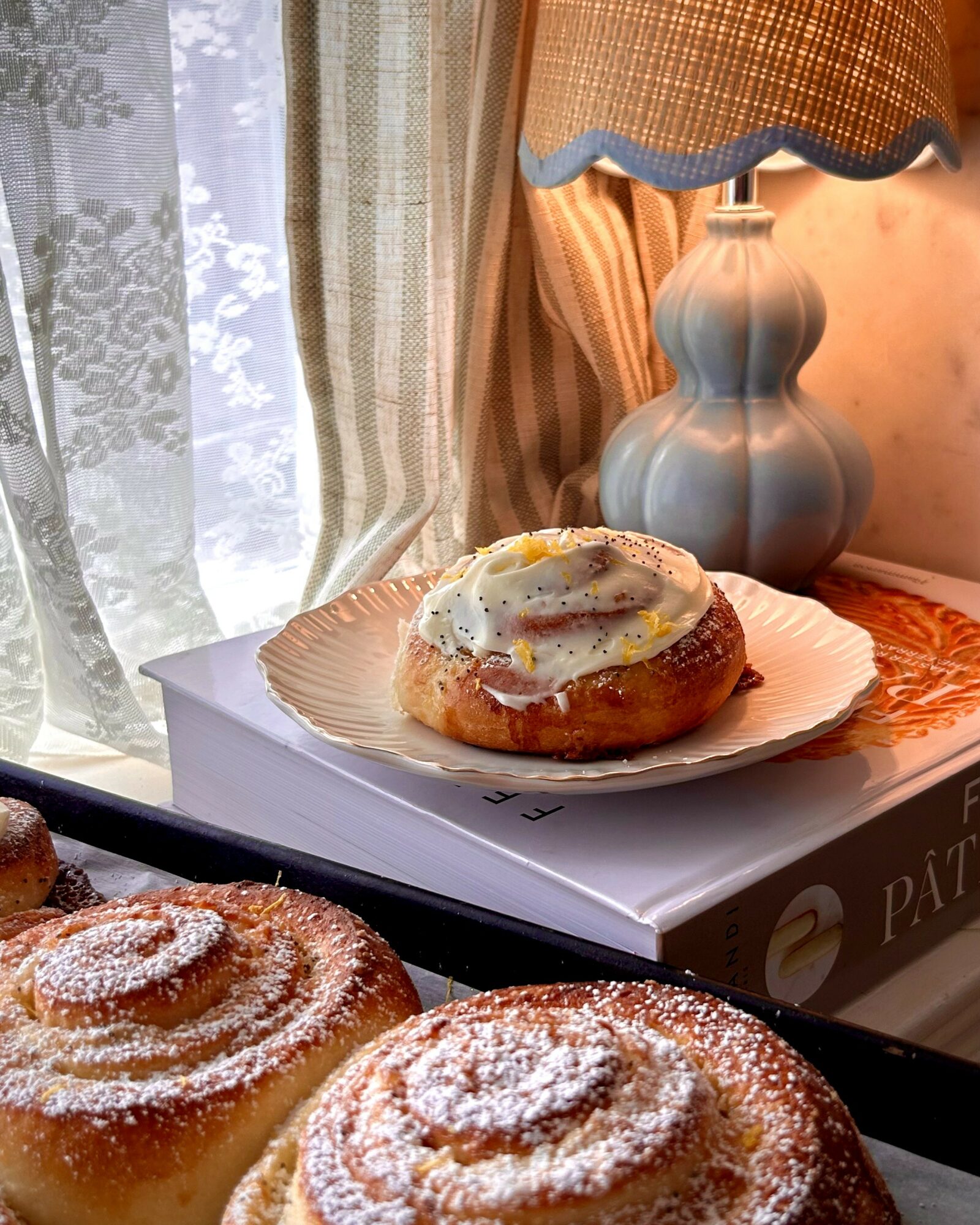 A plate with a pastry topped with icing and sprinkles, placed on a white box near a window with lace curtains, a lamp, and a book.