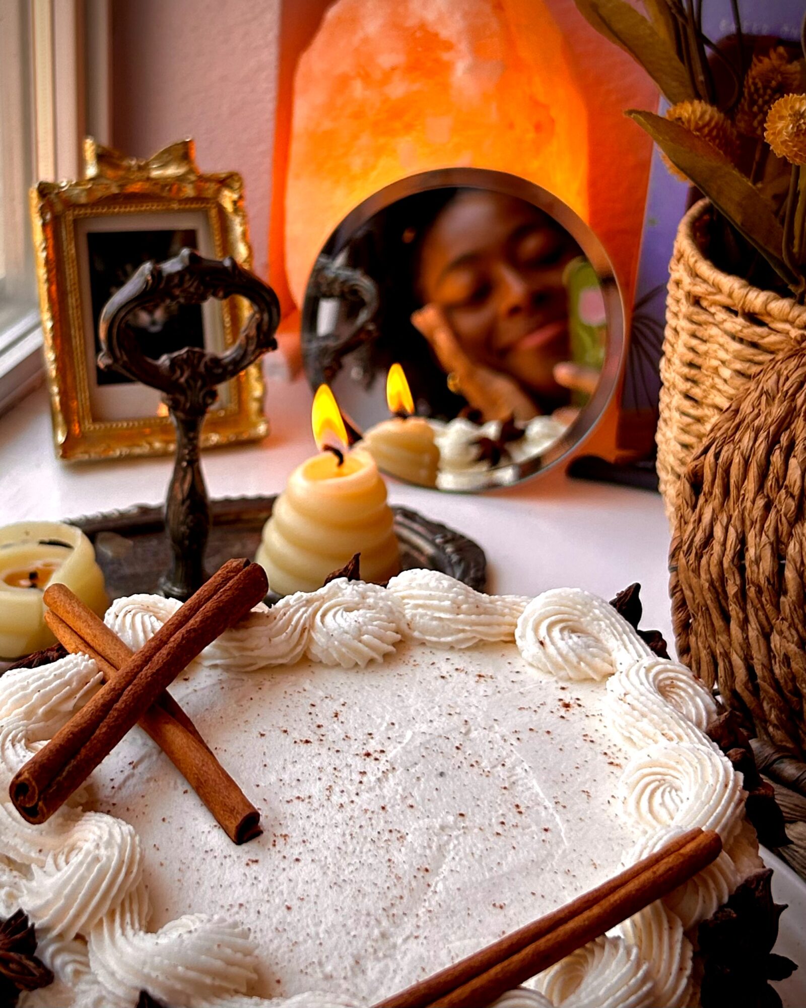 Decorative table with candles, a mirror reflecting a smiling person, framed picture, and a basket with flowers, in warm lighting.