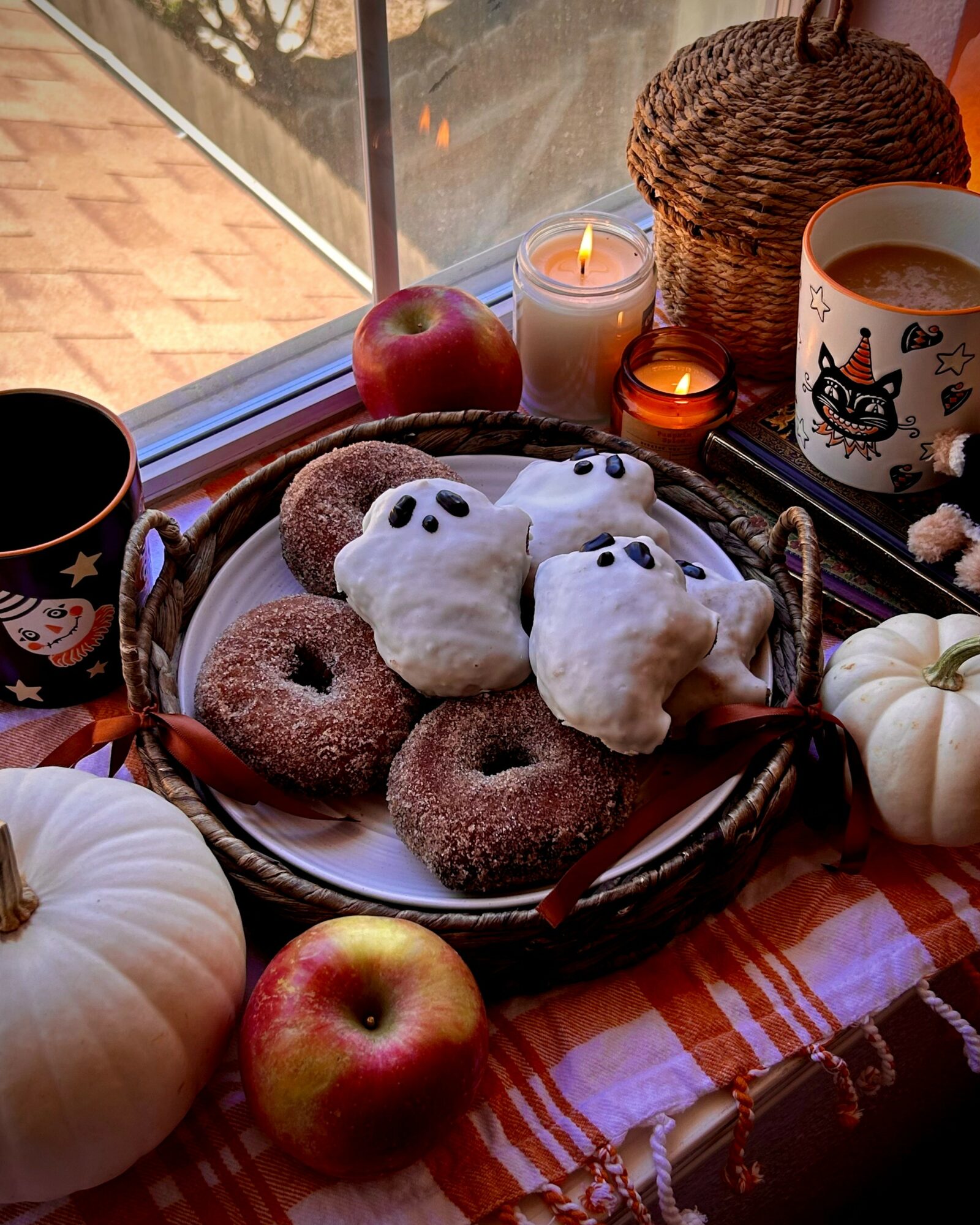 Plate with ghost-shaped cookies and donuts on a table near a window, surrounded by pumpkins, candles, and a mug with a cat design.