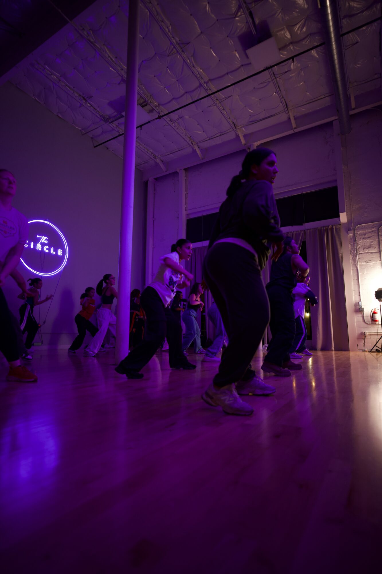People dancing in a dimly lit room with purple lighting and a circular logo on the wall.