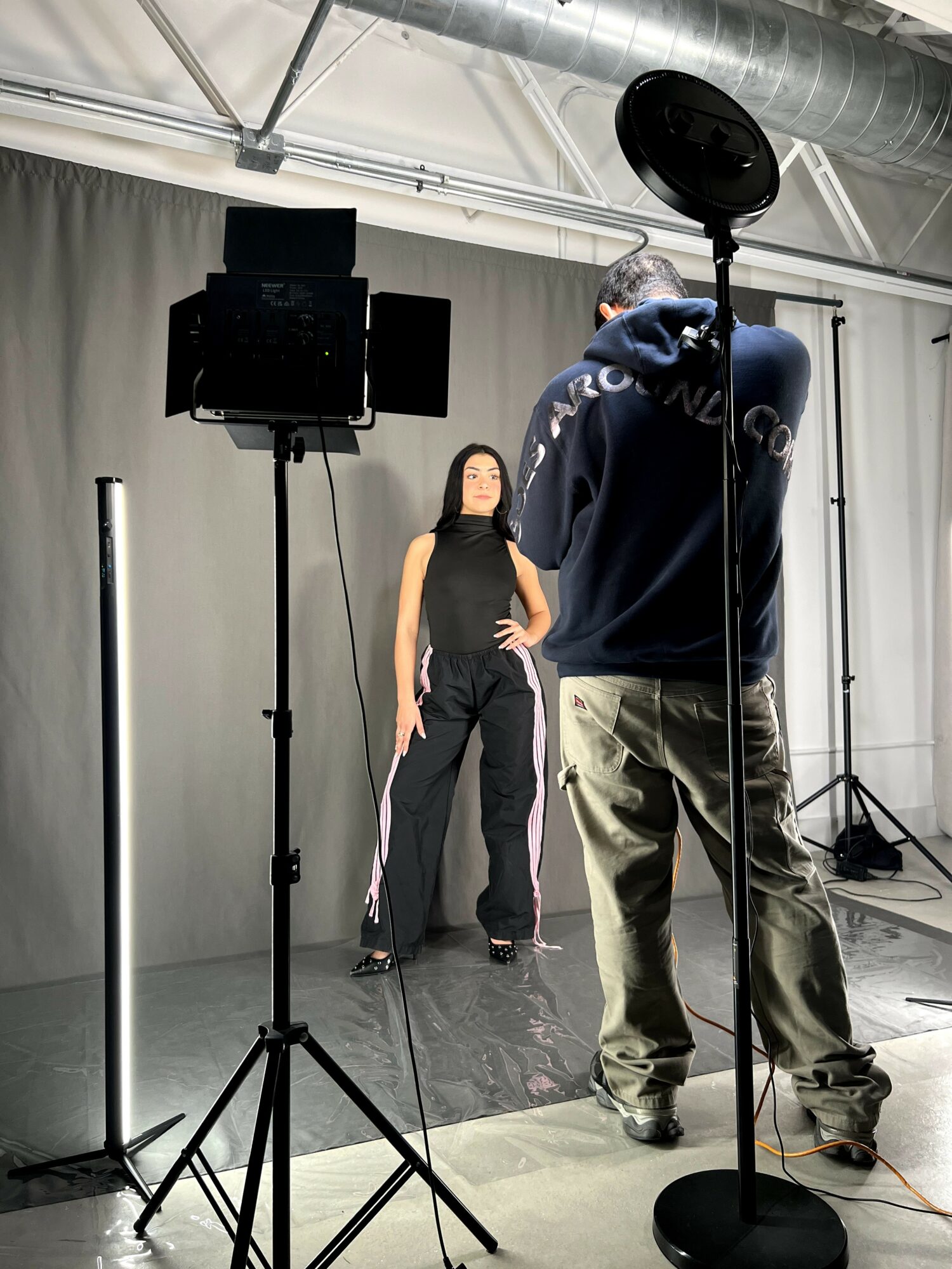 A woman poses on a black stage with a man filming her, in an industrial space with equipment and lighting.