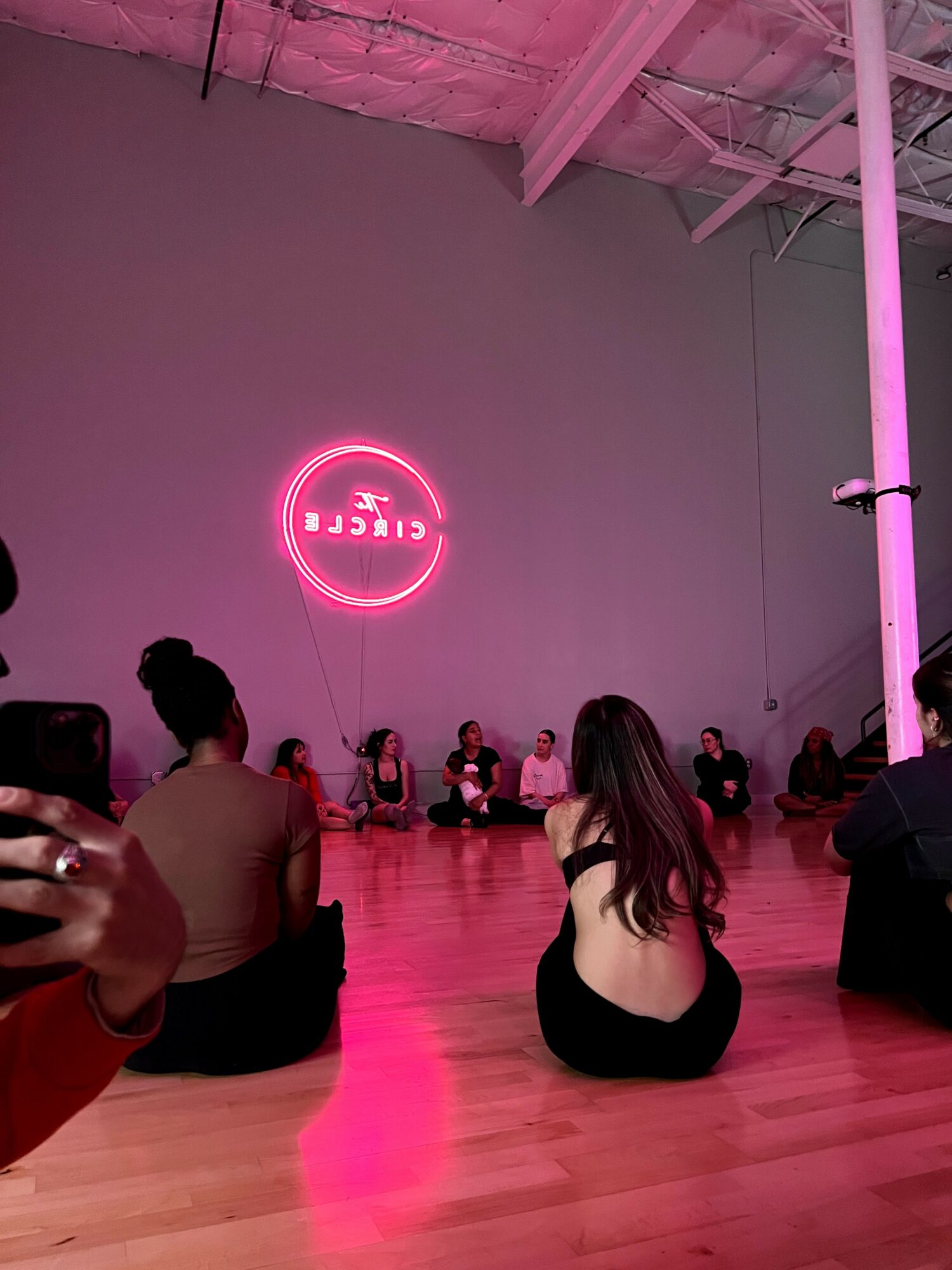 People sitting on a wooden floor in a dimly lit room with a pink neon sign on the wall.