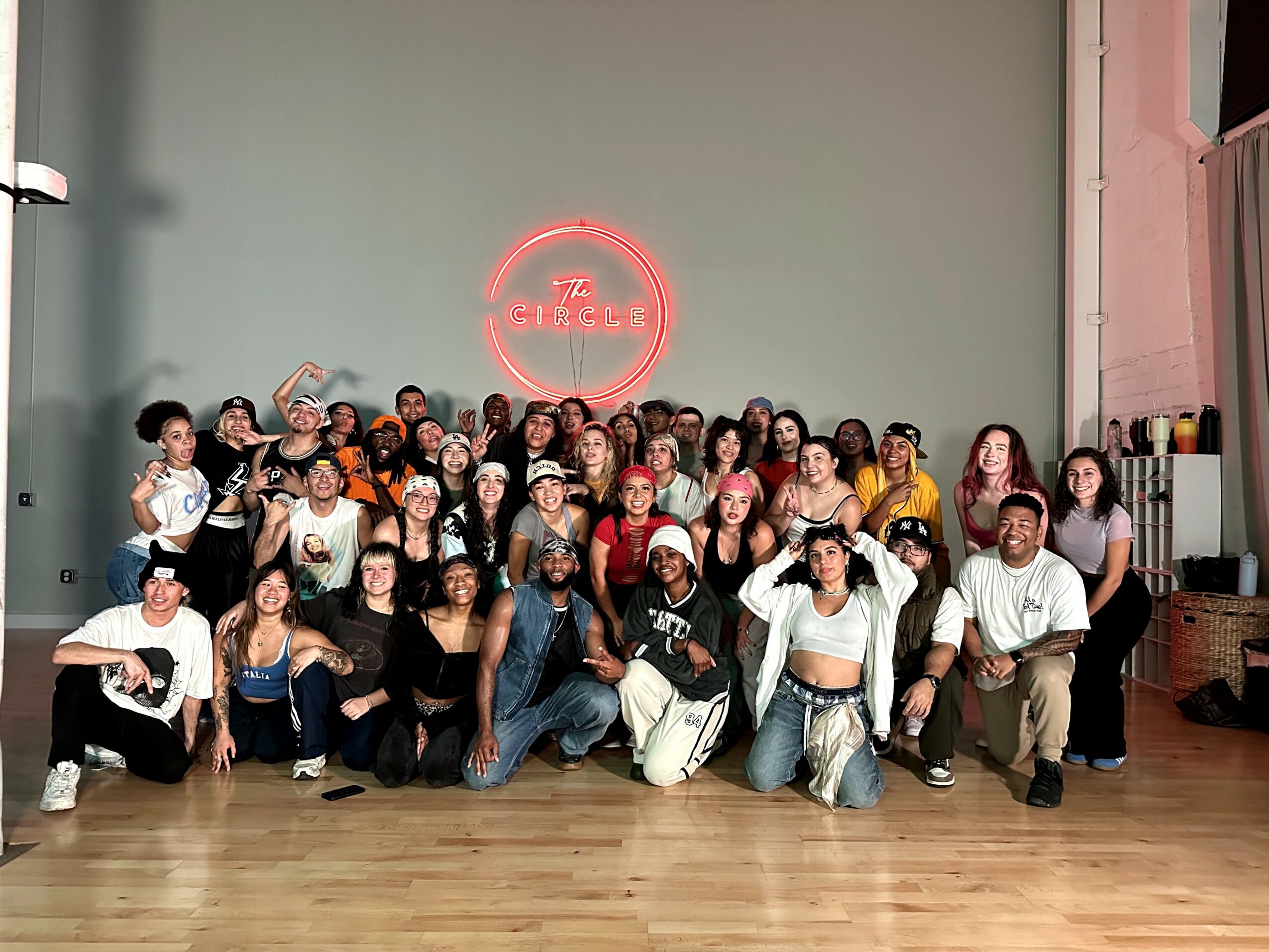 Group of people posing in front of a wall with a neon sign, some making gestures, in a dance studio or event space.
