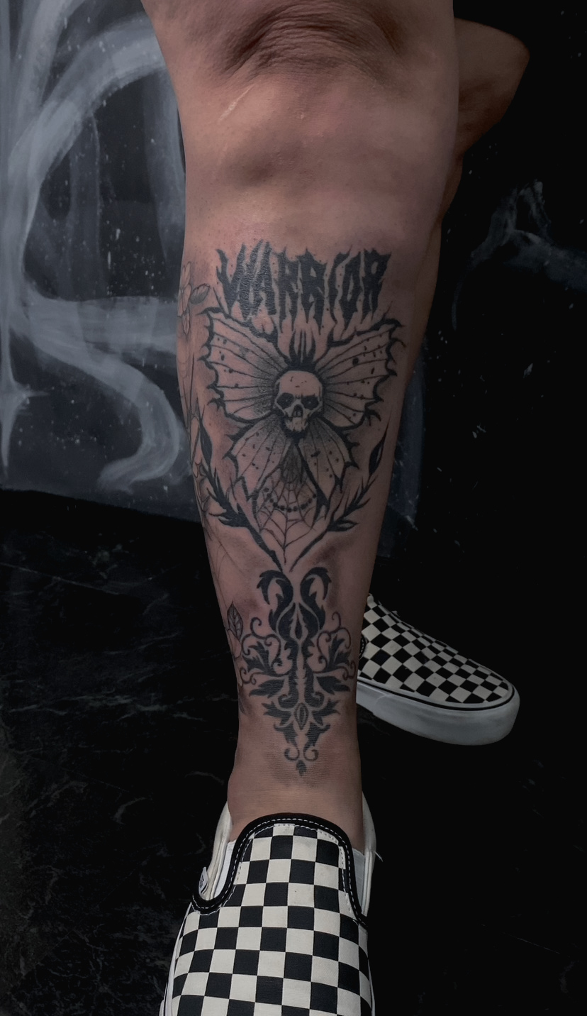 Tattoo of a skull with wings and decorative patterns on a person's lower leg, with checkered shoes nearby.