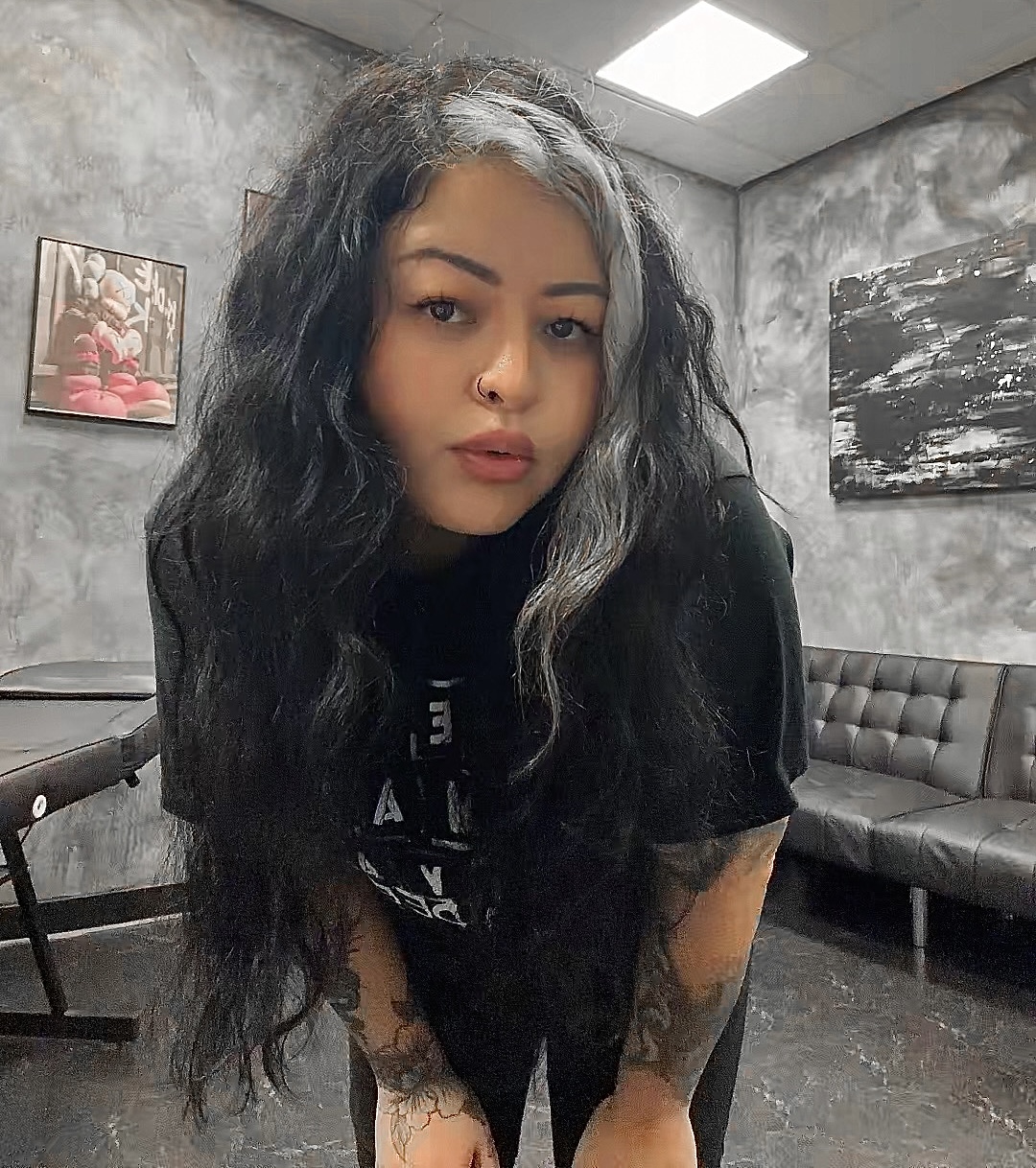 Young woman with long, wavy black hair and tattoos on her arms, standing in a room with gray walls and artwork.
