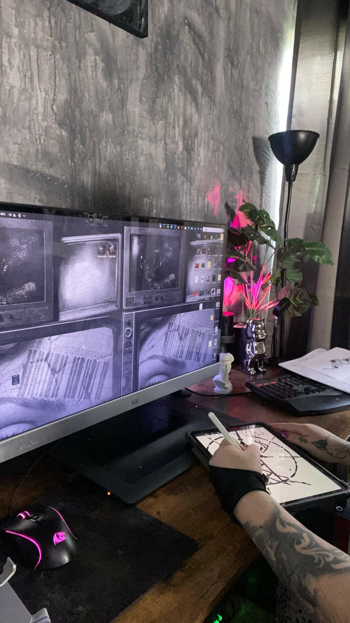 Desk with computer monitor displaying multiple images, a mouse, a hand with a tattoo, and a plant in the background.