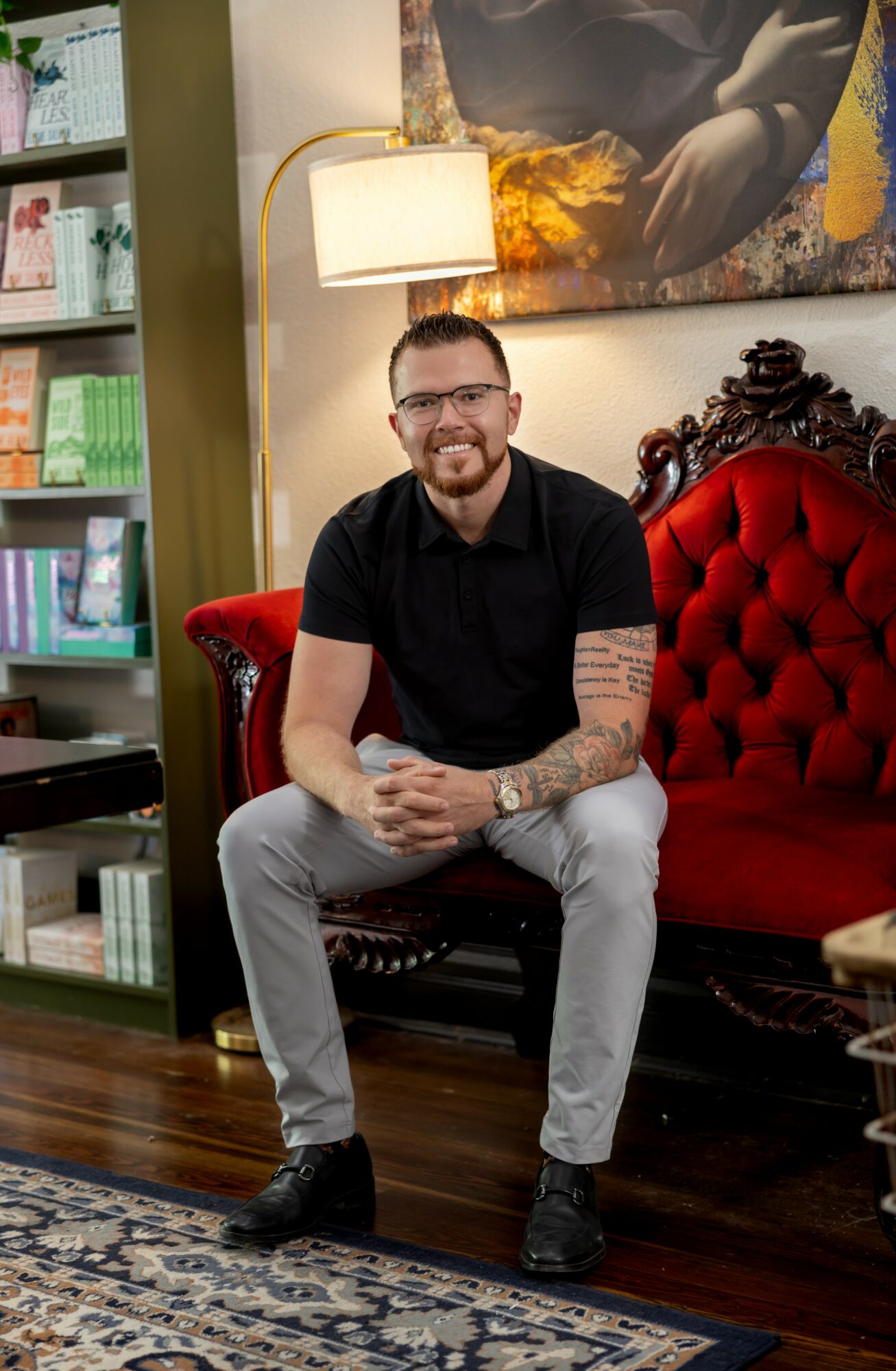 Man with glasses and tattoos sitting on a red velvet sofa in a room with bookshelf and artwork.