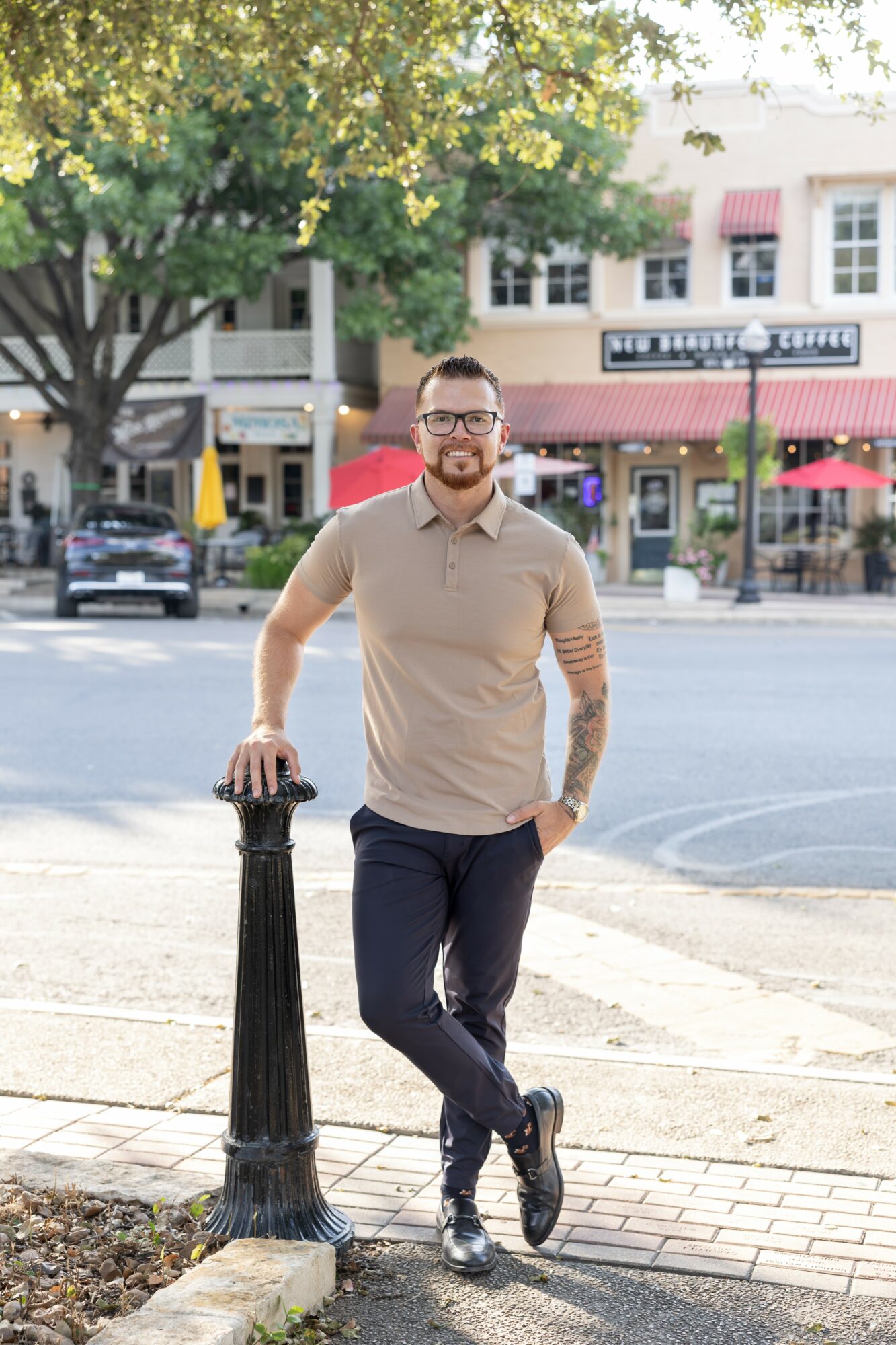 Man with glasses and a beard stands outdoors on a city sidewalk, leaning on a black post, with trees and buildings in background.