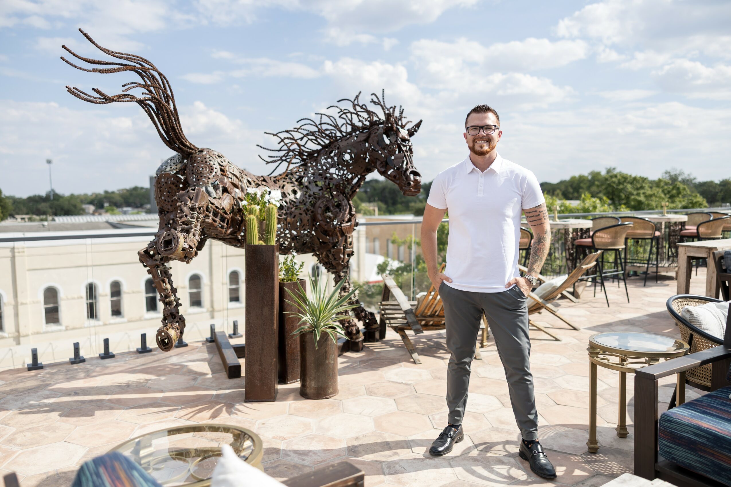Man standing on rooftop terrace with metal horse sculpture, potted plants, and outdoor furniture, cityscape in background.