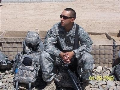 Soldier in camouflage uniform sitting on a bench with a rifle, wearing sunglasses, in a desert-like environment.