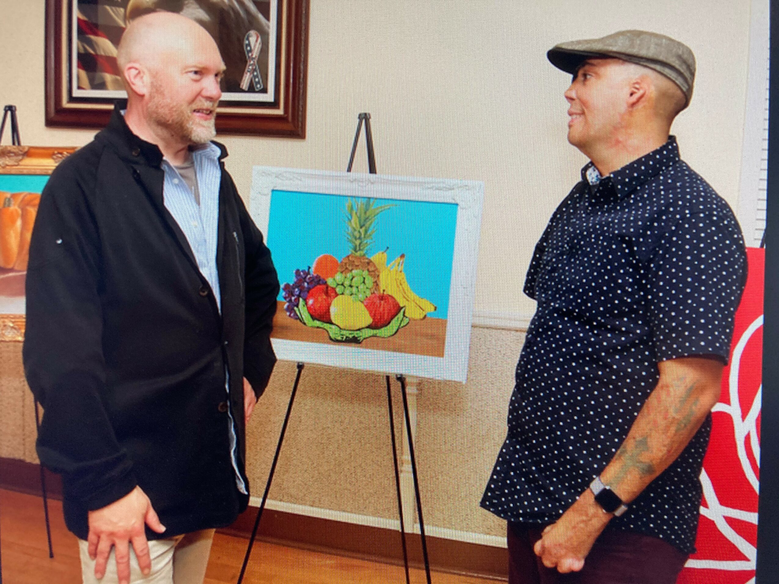 Two men stand facing each other near a colorful fruit painting on an easel, indoors with framed artwork on the wall.