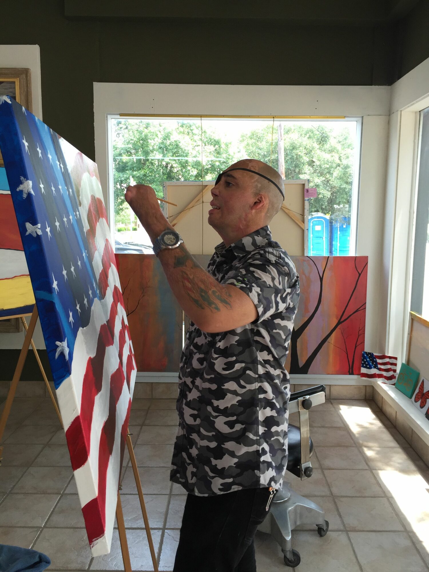Man with tattoos and glasses painting an American flag on a canvas indoors, with a window and trees outside.