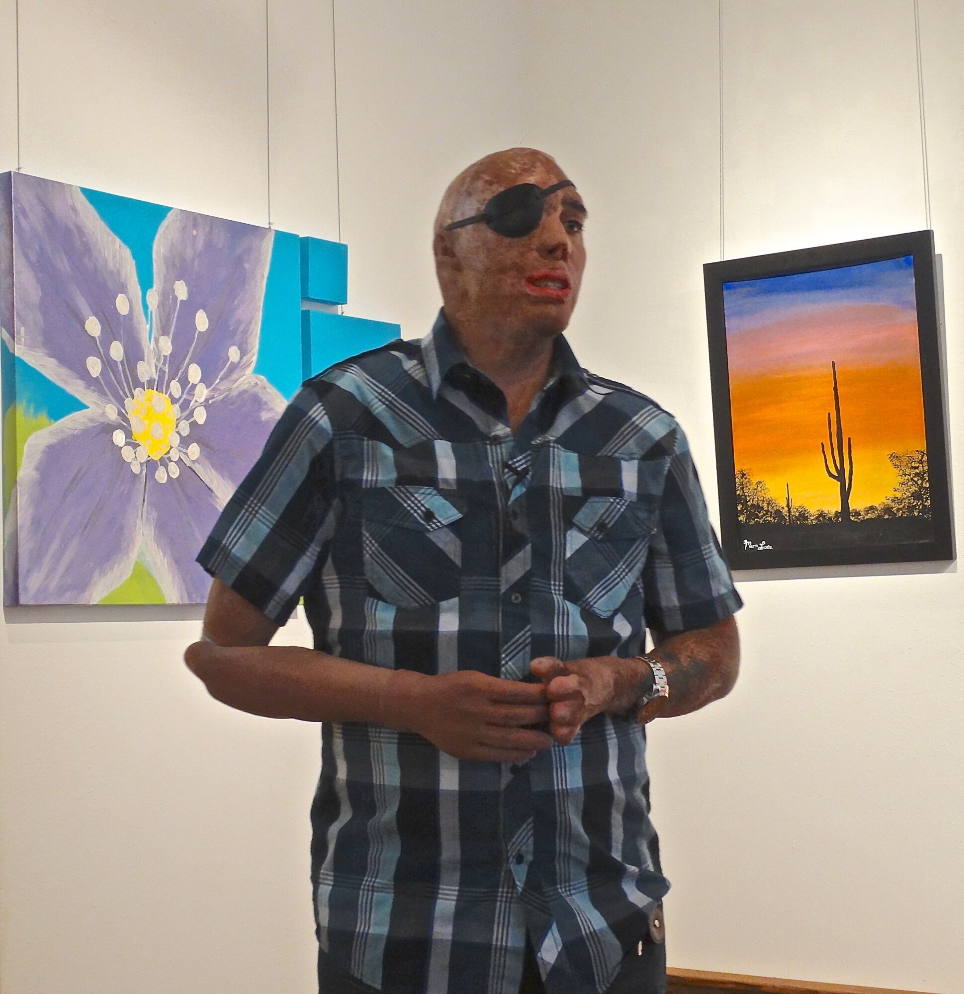 Person with sunglasses wearing a plaid shirt, standing in front of colorful artwork and a desert landscape painting.