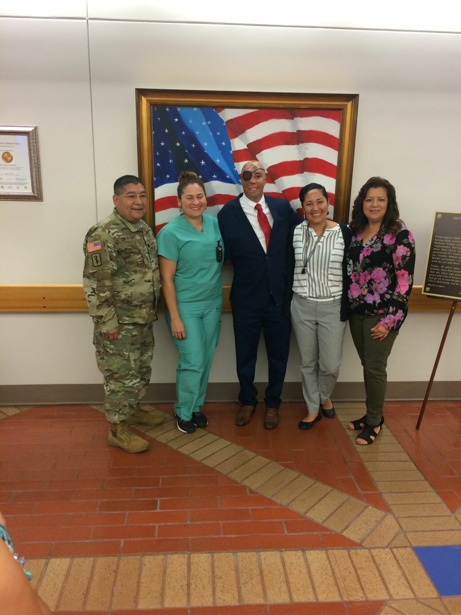 Group of five people standing in front of a framed American flag, indoors, with a brick floor and white wall.