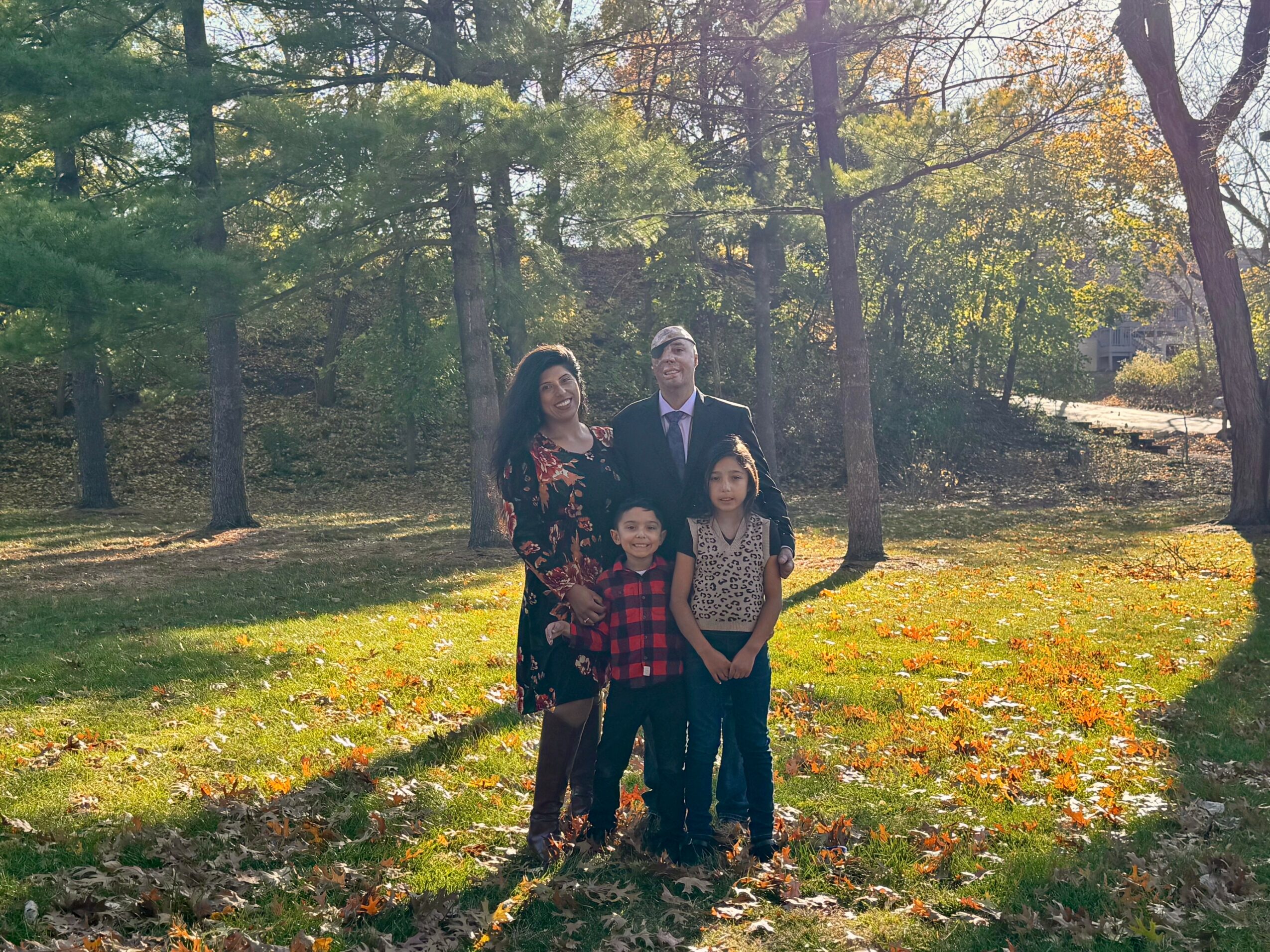 Family of four standing outdoors in a park with trees and fallen leaves, sunny day, group includes two adults and two children.