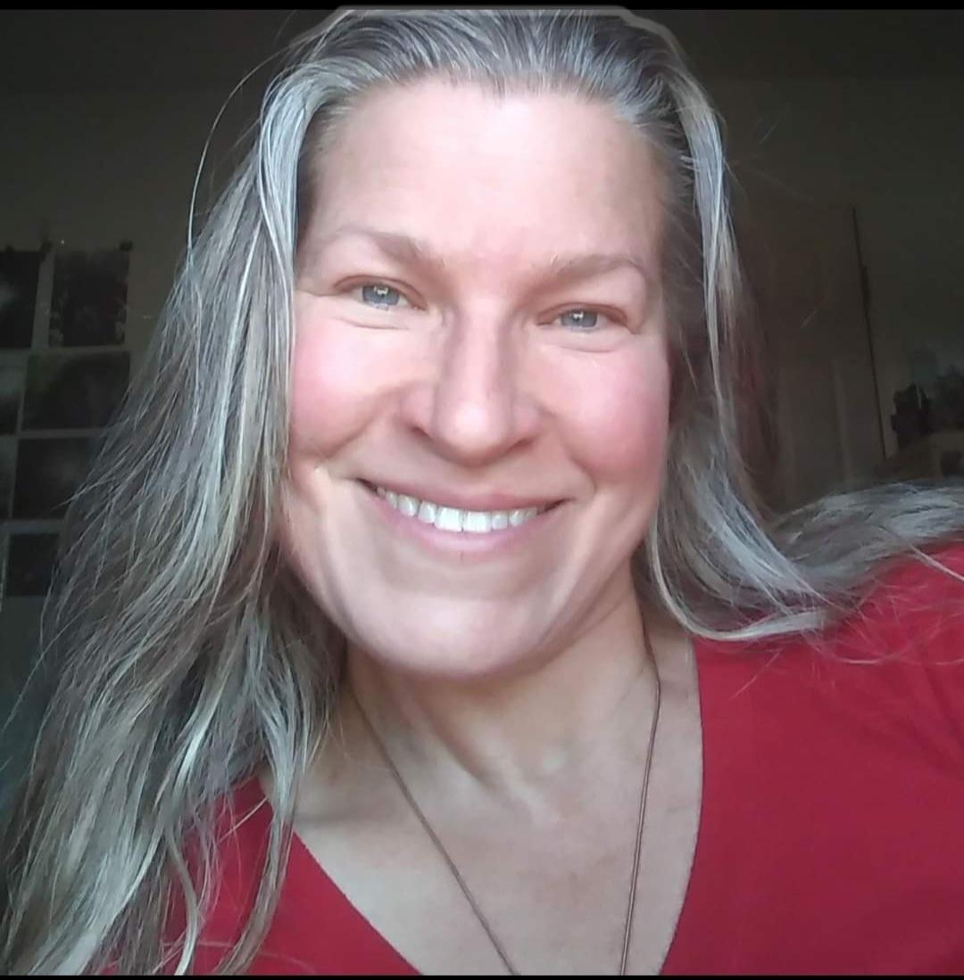 Smiling woman with long gray hair wearing a red top, indoors, natural light, neutral background.