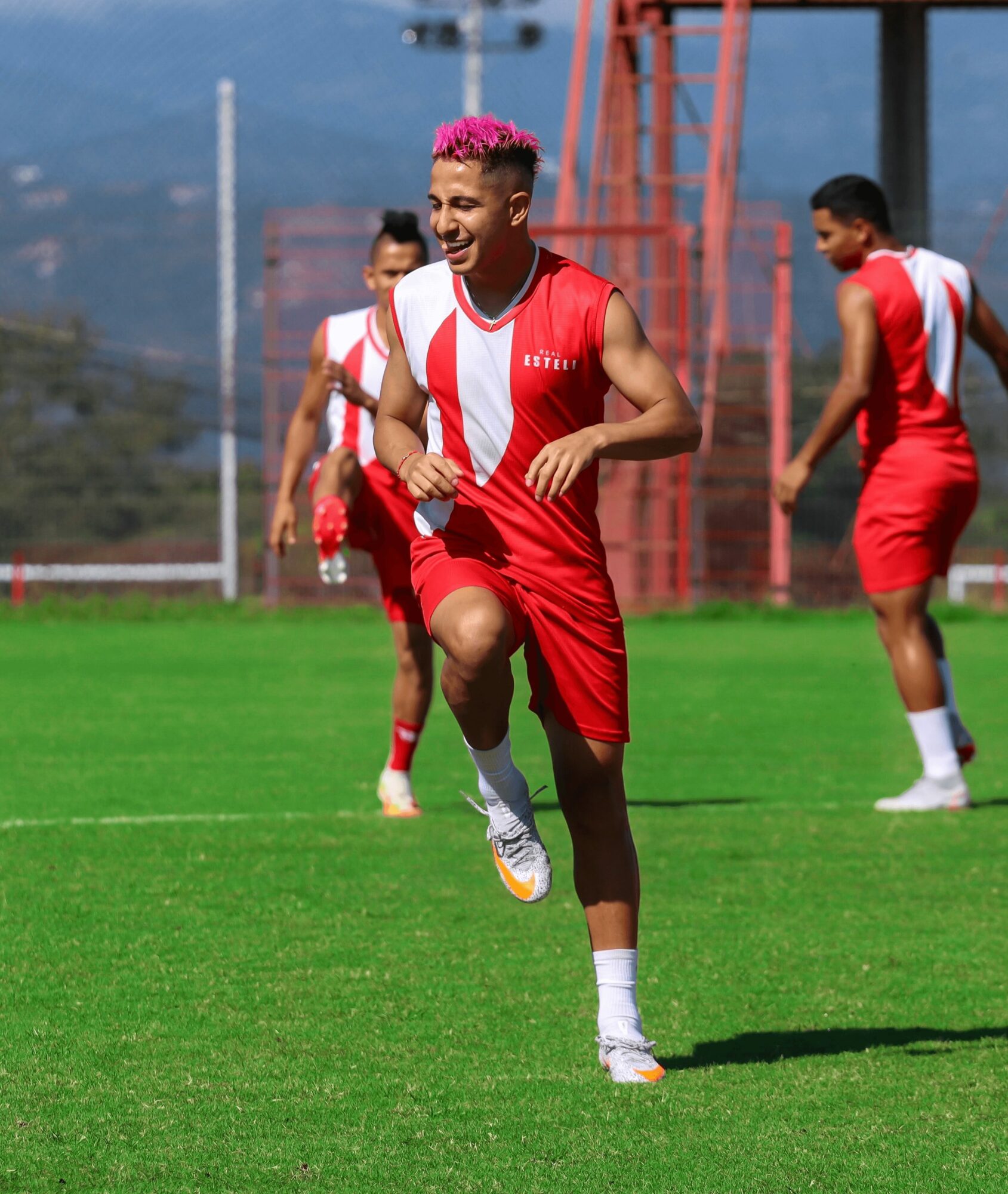 Young male athlete with pink hair running on a soccer field, wearing a red and white uniform, with other players in the background.