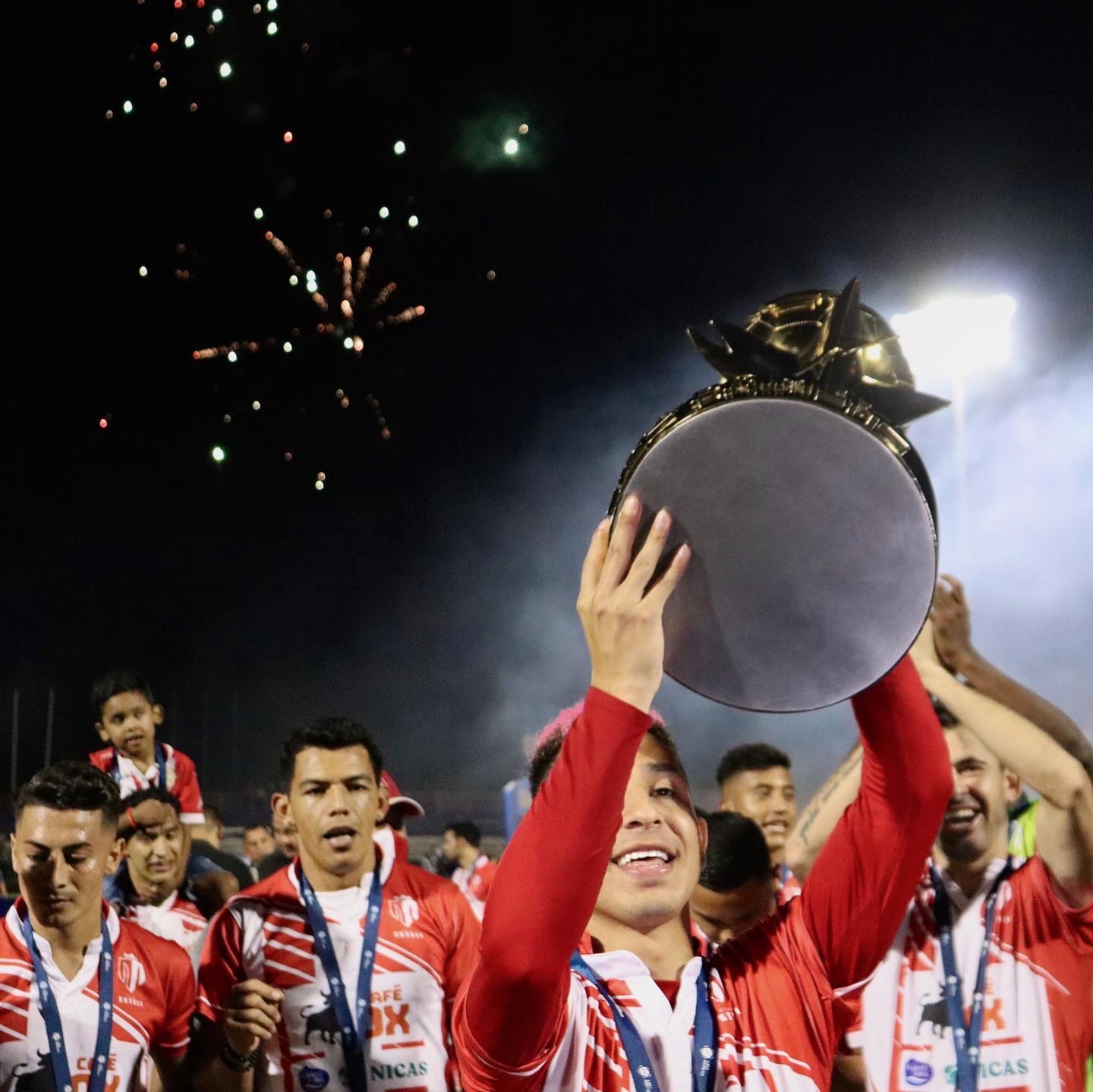 Person holding a trophy above their head with fireworks in the night sky, surrounded by celebrating people in red and white uniforms.