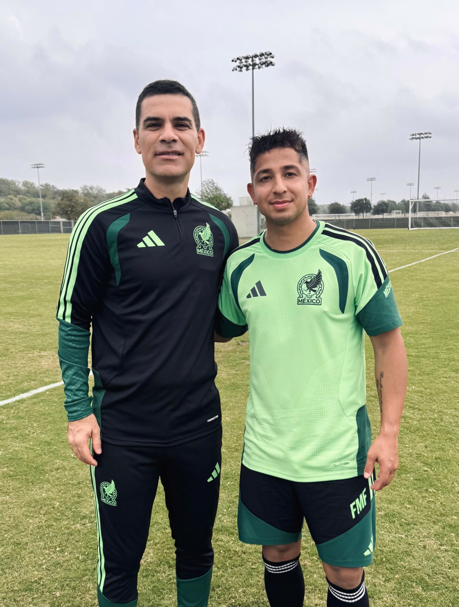 Two men in sportswear standing on a grassy field with light poles in the background.