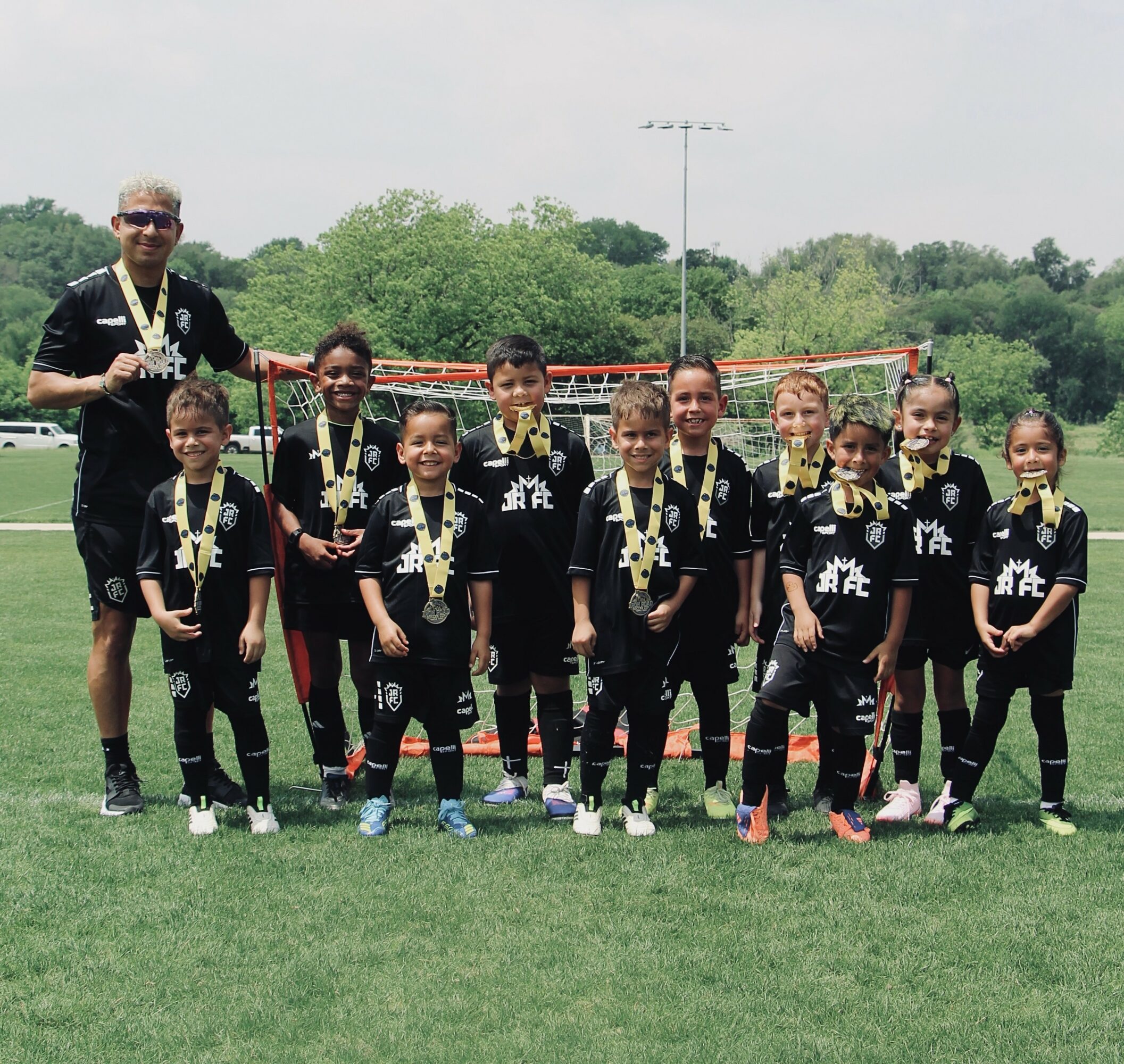 Group of young children in soccer uniforms with medals, standing on a field with a coach, trees, and a goal in the background.