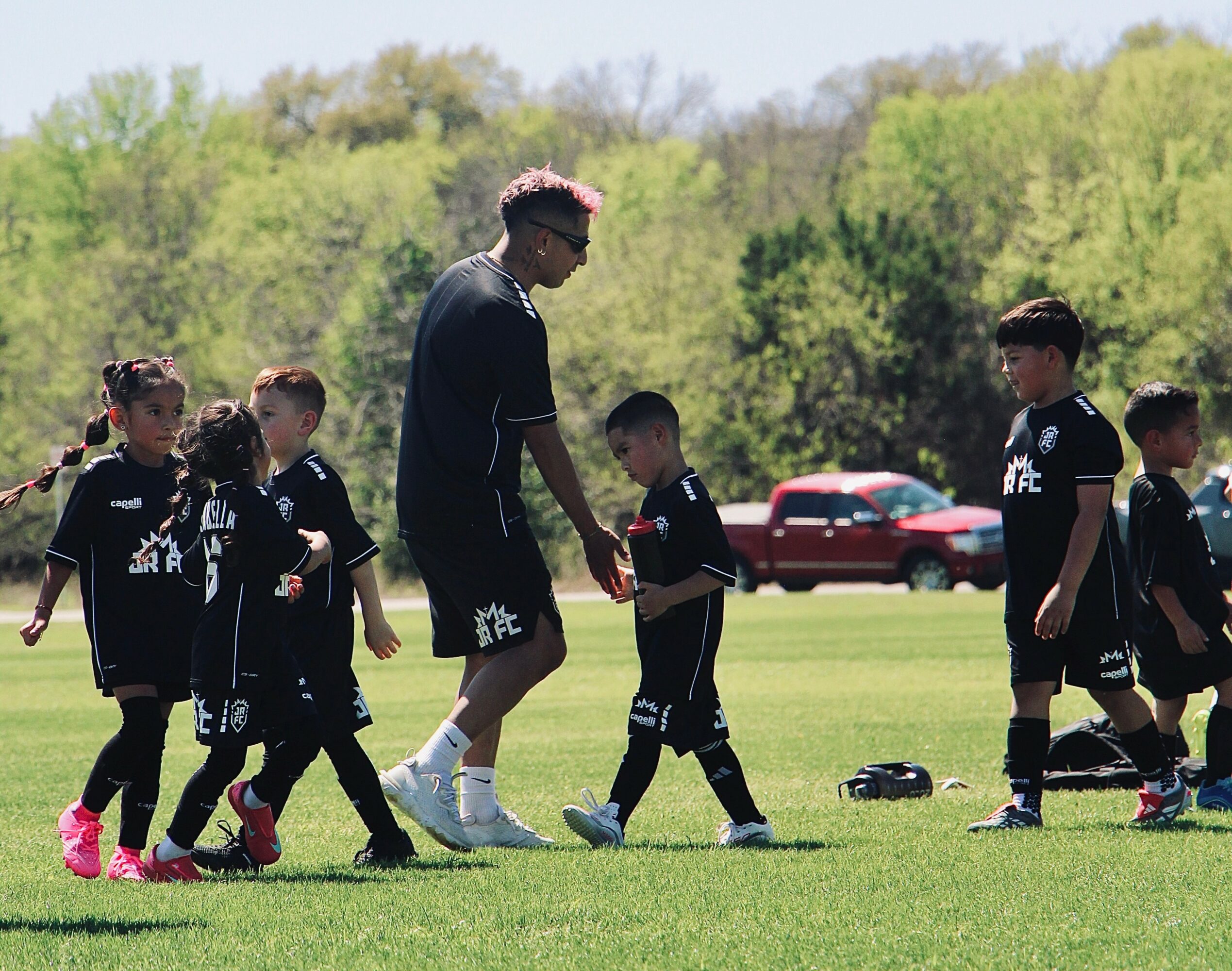 Coach with children on a soccer field, trees and cars in background, sunny day.