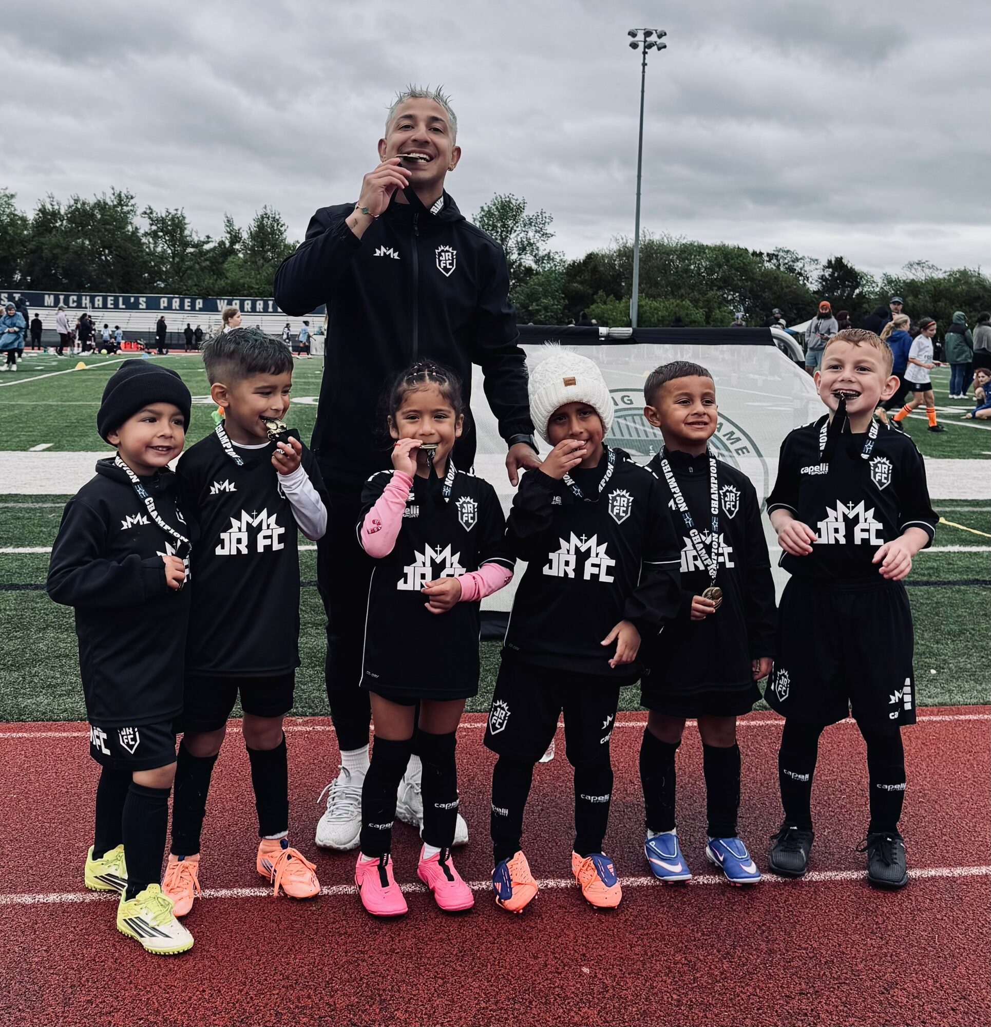 Group of young children in black sports uniforms with a coach on a track field, smiling and holding drinks.