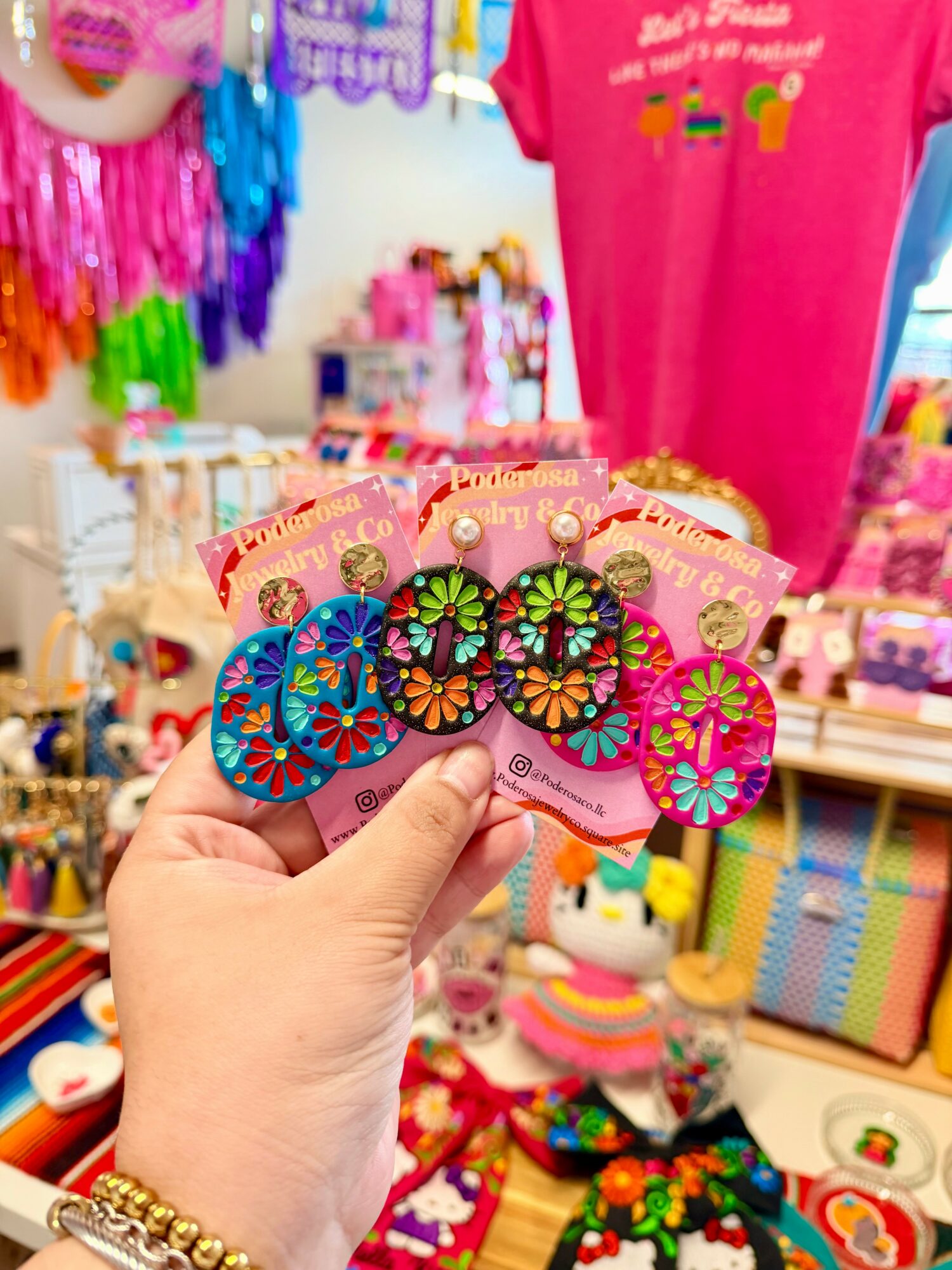Hand holding colorful flower-shaped earrings in a store with various toys and accessories in the background.