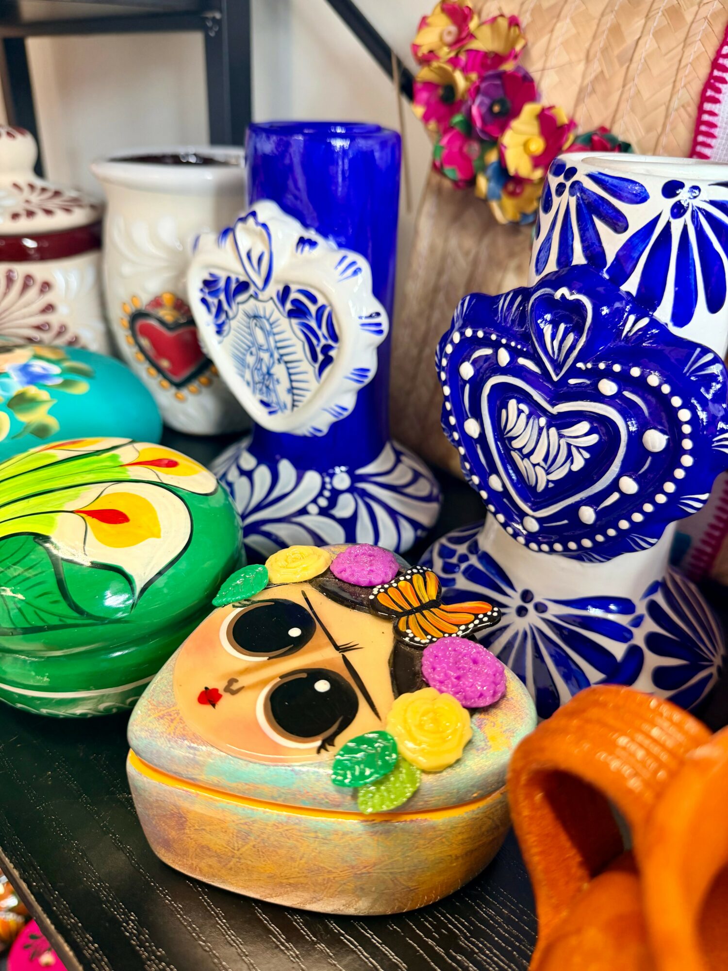 Colorful decorated cake with eyes, flowers, and candies, surrounded by vibrant pottery and flowers.