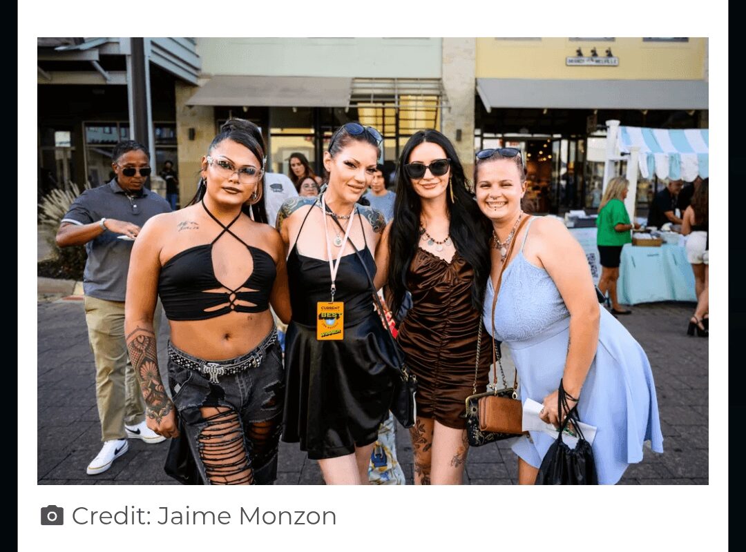 Four women standing together outdoors, smiling, with buildings and people in the background.