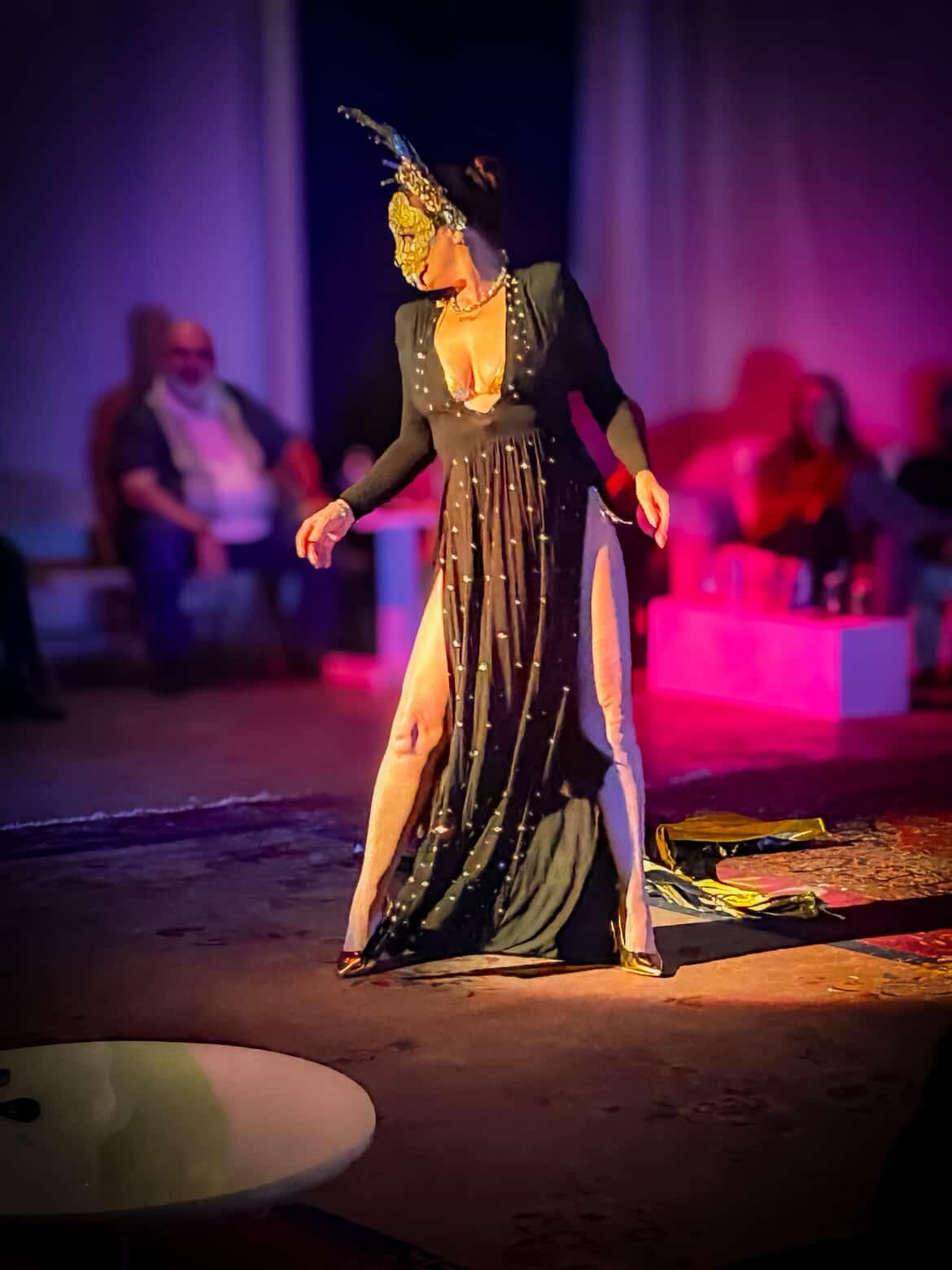 Performer in a black and gold costume with a headdress, dancing on stage with seated audience in background.