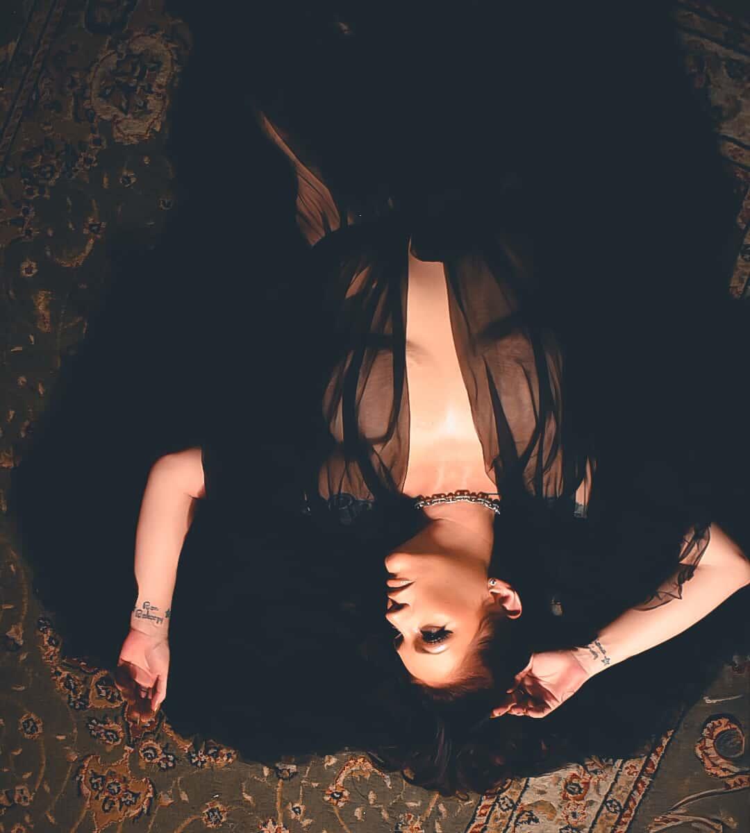 Woman lying on a patterned rug, wearing a black dress with sheer fabric, with arms outstretched and eyes closed.
