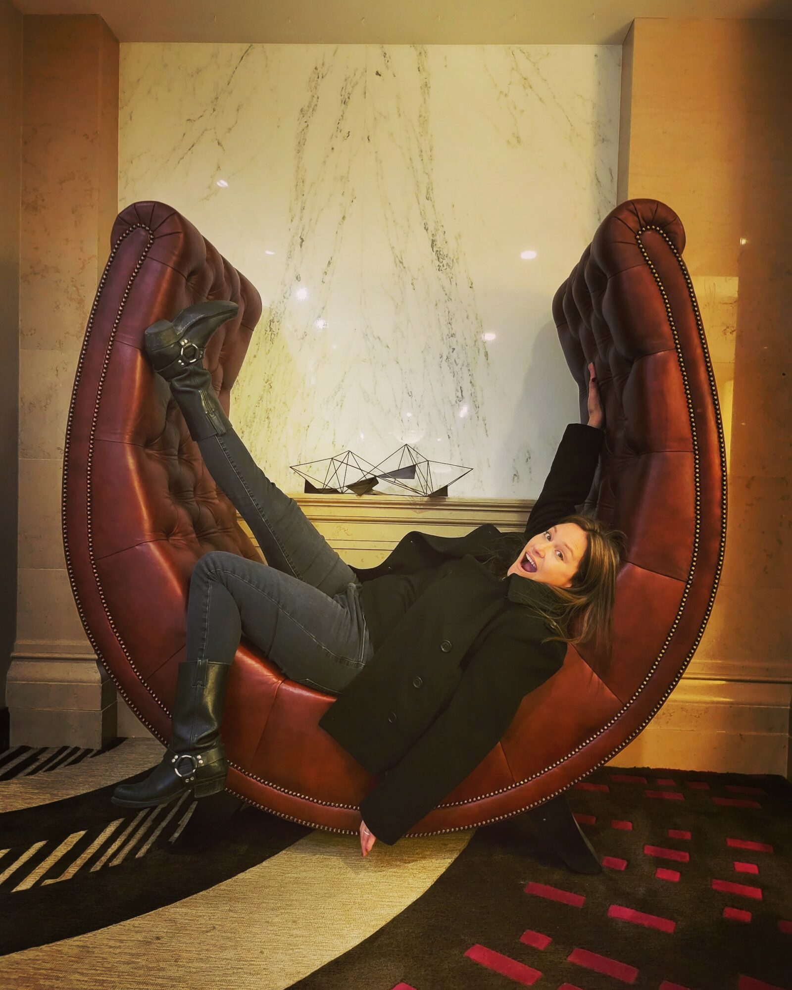 Person lying on a large, curved, red chair with high backrests in a room with marble wall and wooden panels.