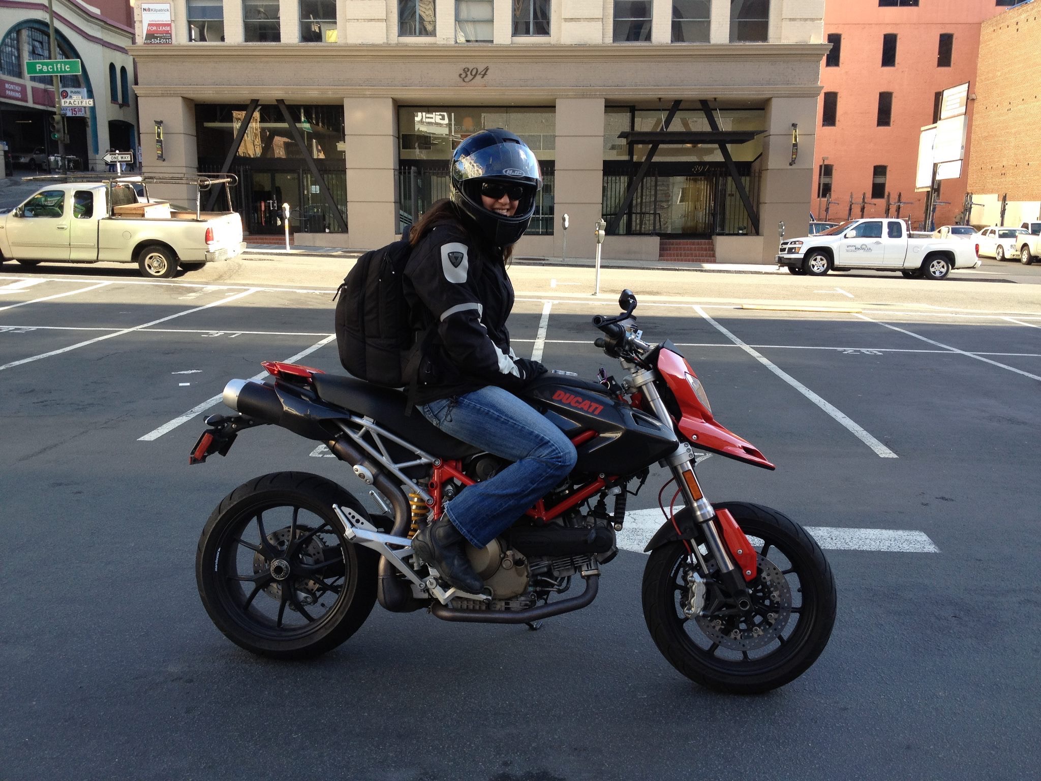 Person wearing a helmet and backpack riding a motorcycle on a city street, buildings in background.