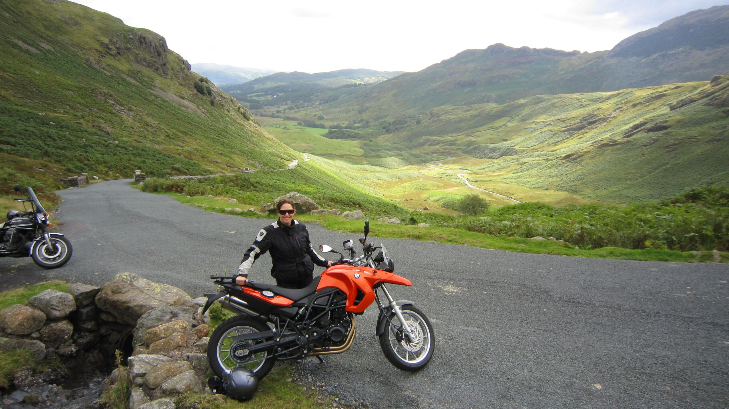 Person in black jacket and helmet sitting on an orange motorcycle on a curved road with green hills and mountains in the background.