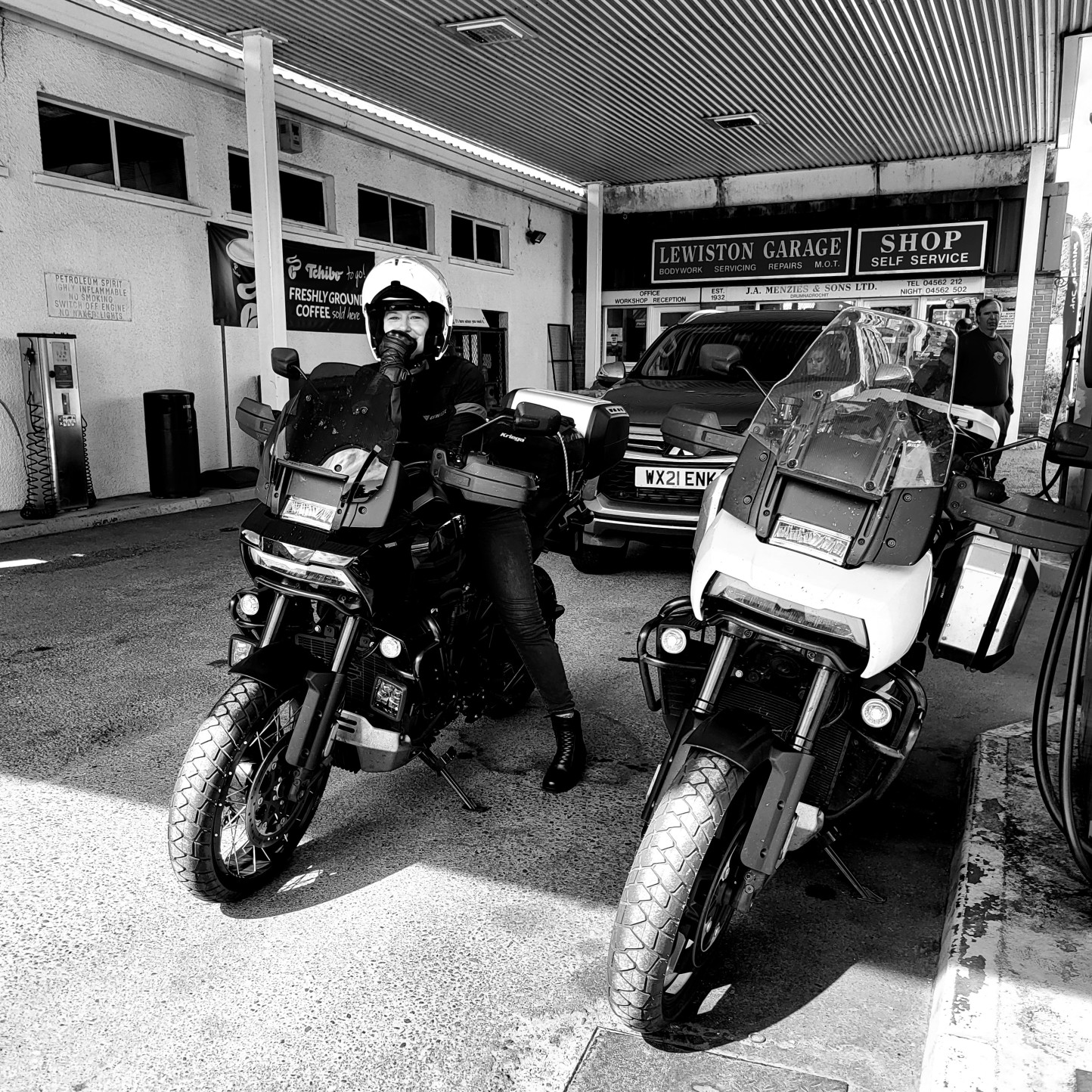 Person wearing helmet and gear sitting on motorcycle at garage, with another motorcycle nearby and cars in background.