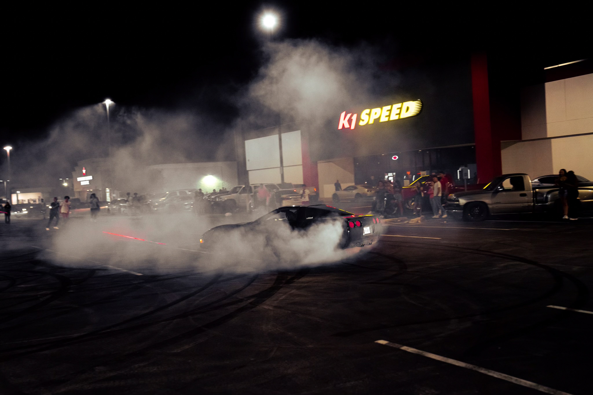Car emits smoke or fire at night outside a store with a sign reading 'KI SPEED,' surrounded by people and parked cars.