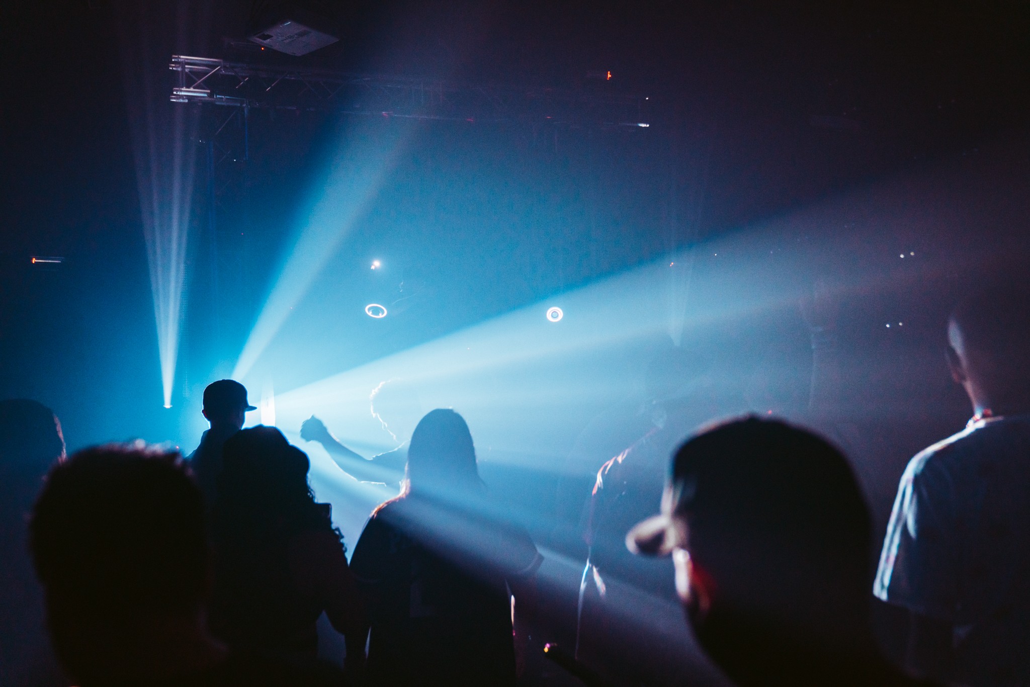 People in a dark room with bright stage lights and beams, some silhouettes visible, possibly at a concert or event.