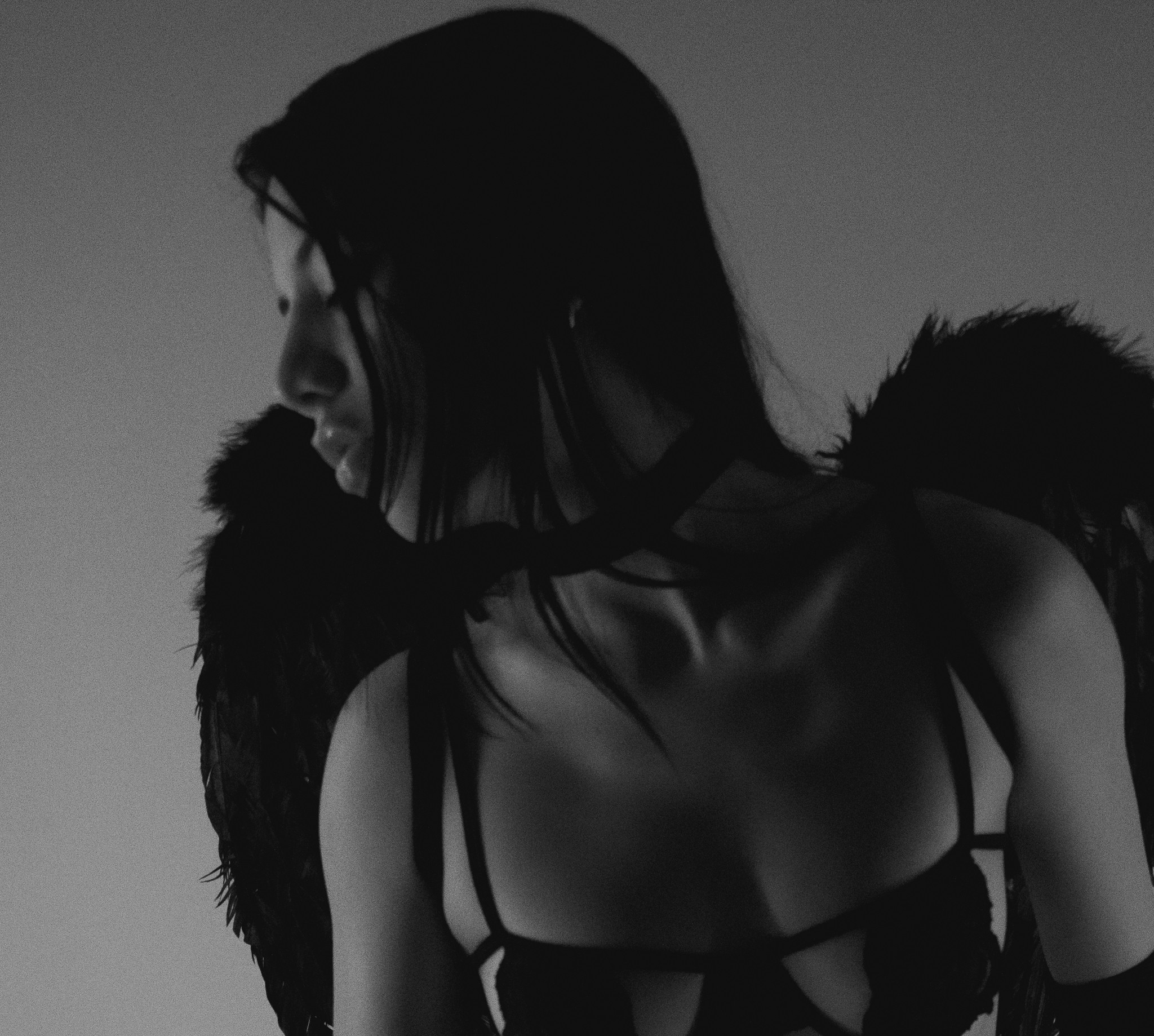Side profile of a woman with dark hair, wearing a strappy top and black feathered wings, in black and white.