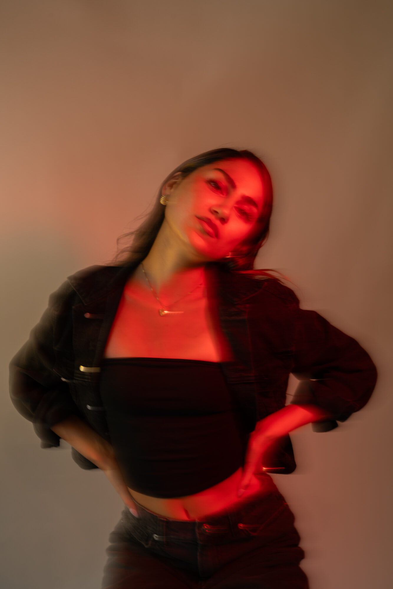 Young woman with long hair, wearing a black top and jacket, posing with one hand on her hip, illuminated by red and warm lighting.