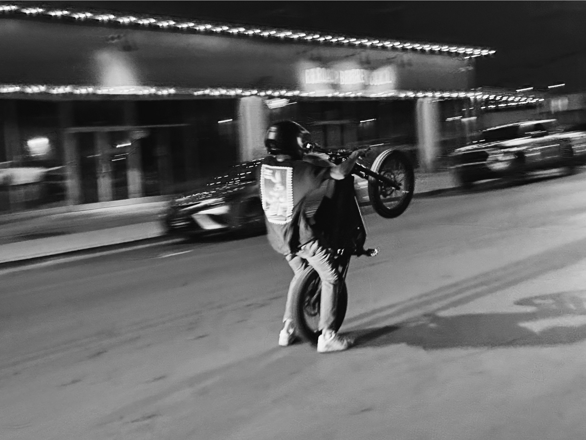 Person wearing a helmet and backpack lifting a bicycle in a city street at night, blurred background of cars and buildings.