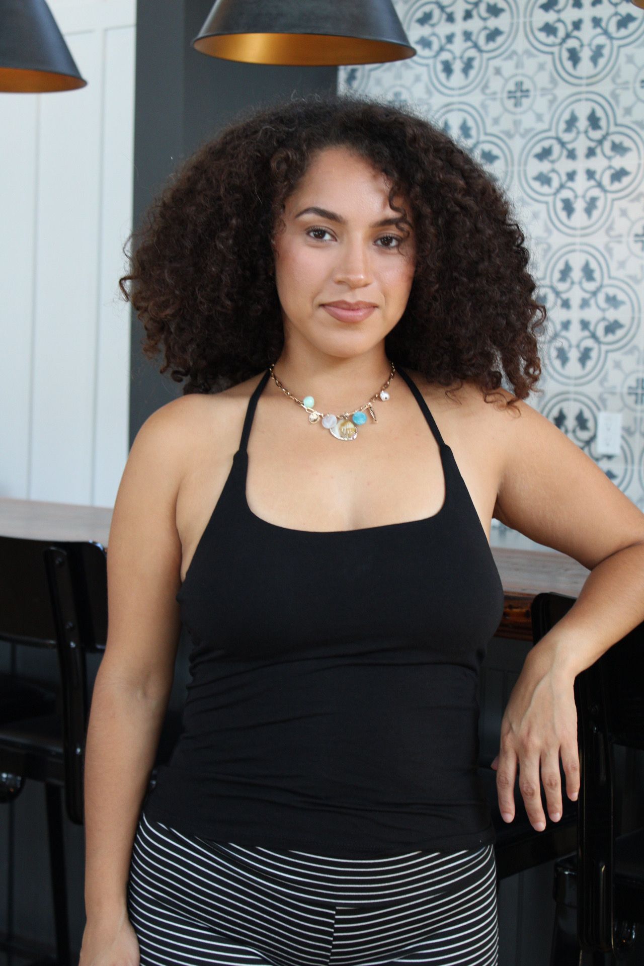 Woman with curly hair wearing a black top and striped skirt, standing indoors with a patterned wall in the background.