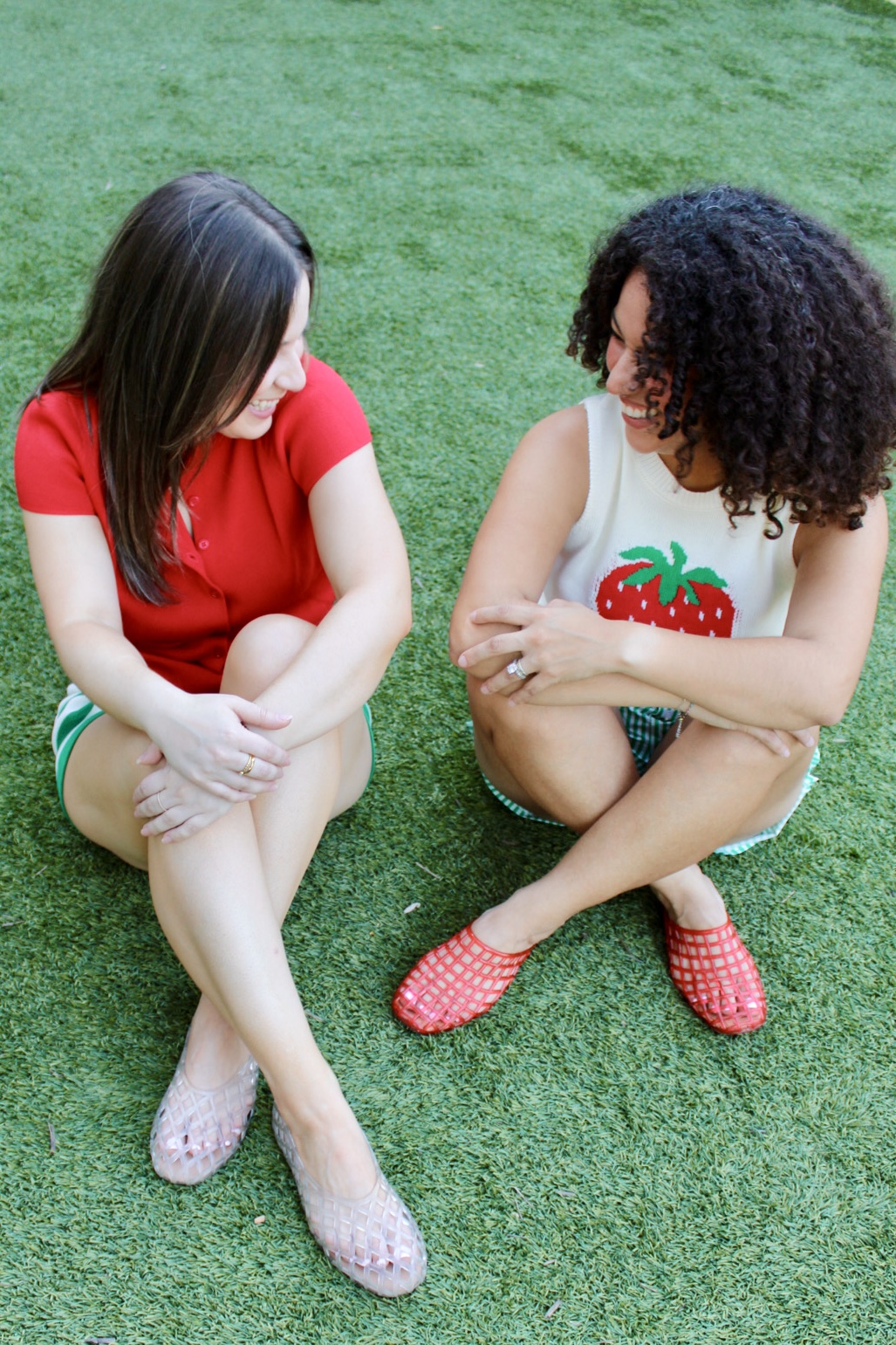 Two women sitting on grass, facing each other, with crossed arms and legs, engaged in conversation, outdoors.