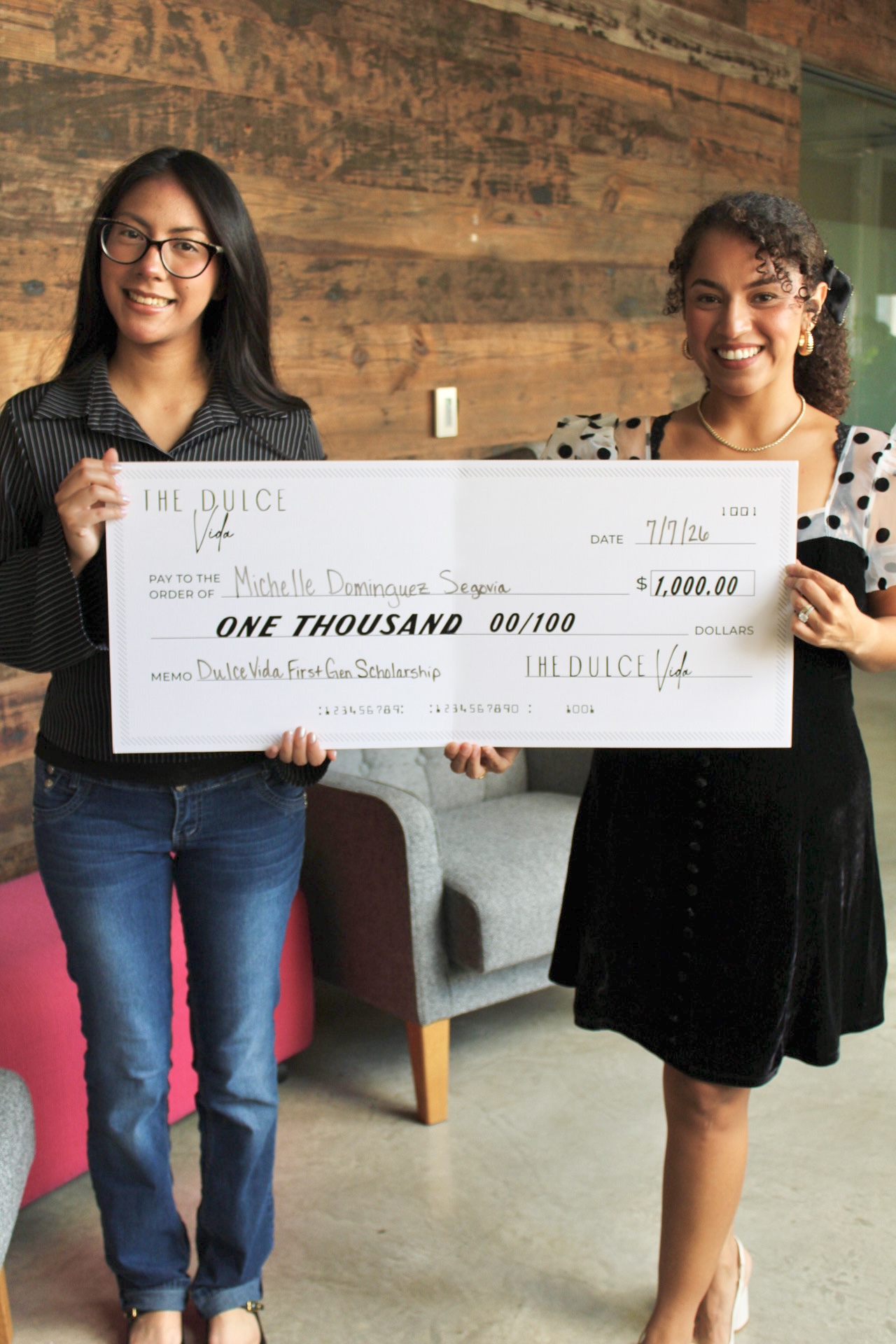 Two women holding a large check, smiling, standing indoors with a wooden wall background.