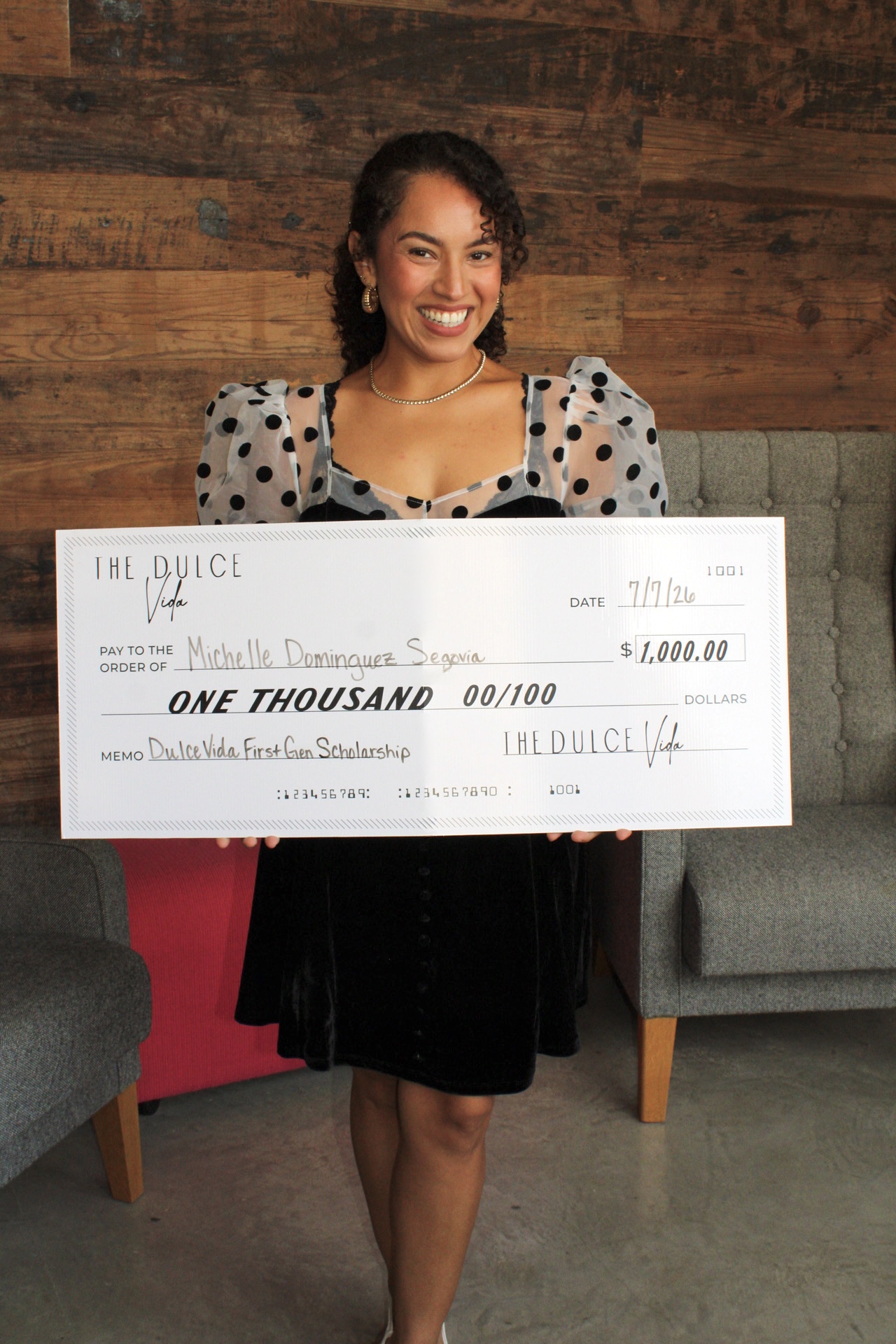 Woman holding oversized check, smiling, standing indoors with wooden wall background, wearing a black dress and polka dot sleeves.