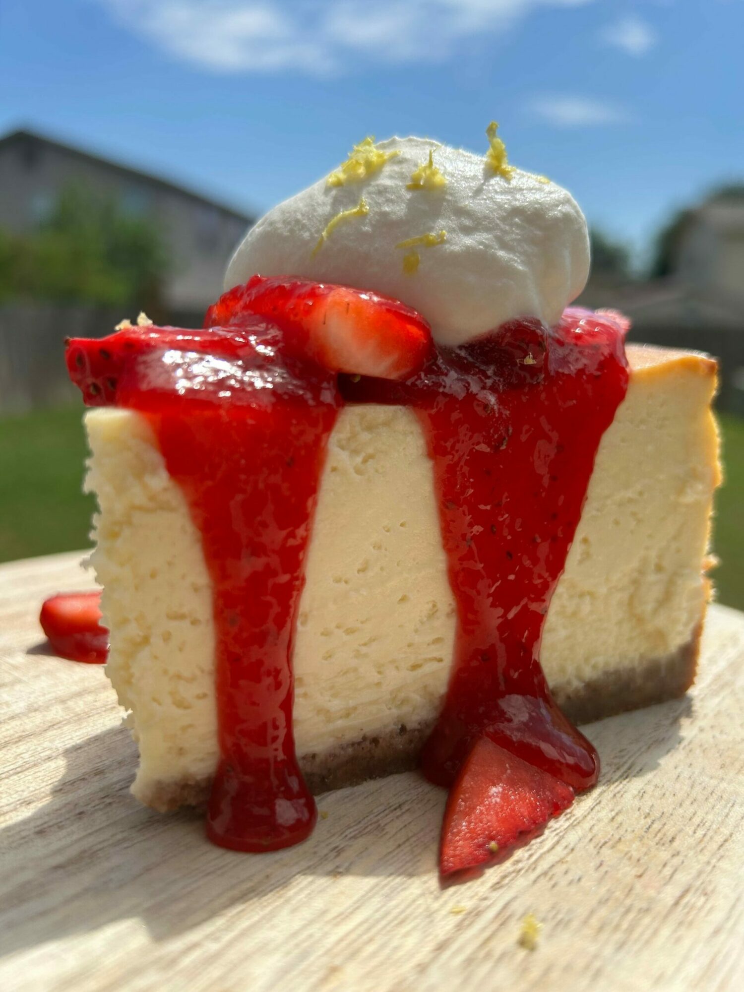 Slice of cheesecake with strawberry sauce, fresh strawberries, and a dollop of whipped cream on top, on a wooden surface.